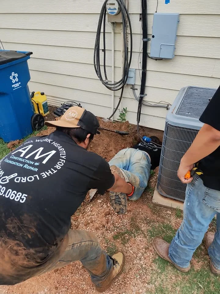 Two workers fix a leak near an air conditioning unit outside a house, with one worker crouching and the other standing nearby.