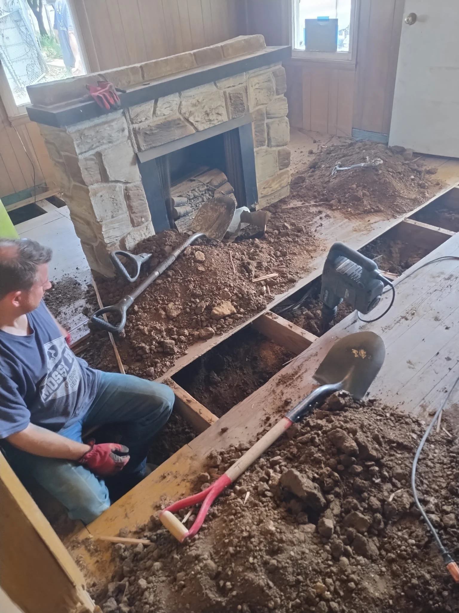 A person working on a home renovation project, digging in a trench in front of a stone fireplace with tools and dirt around.