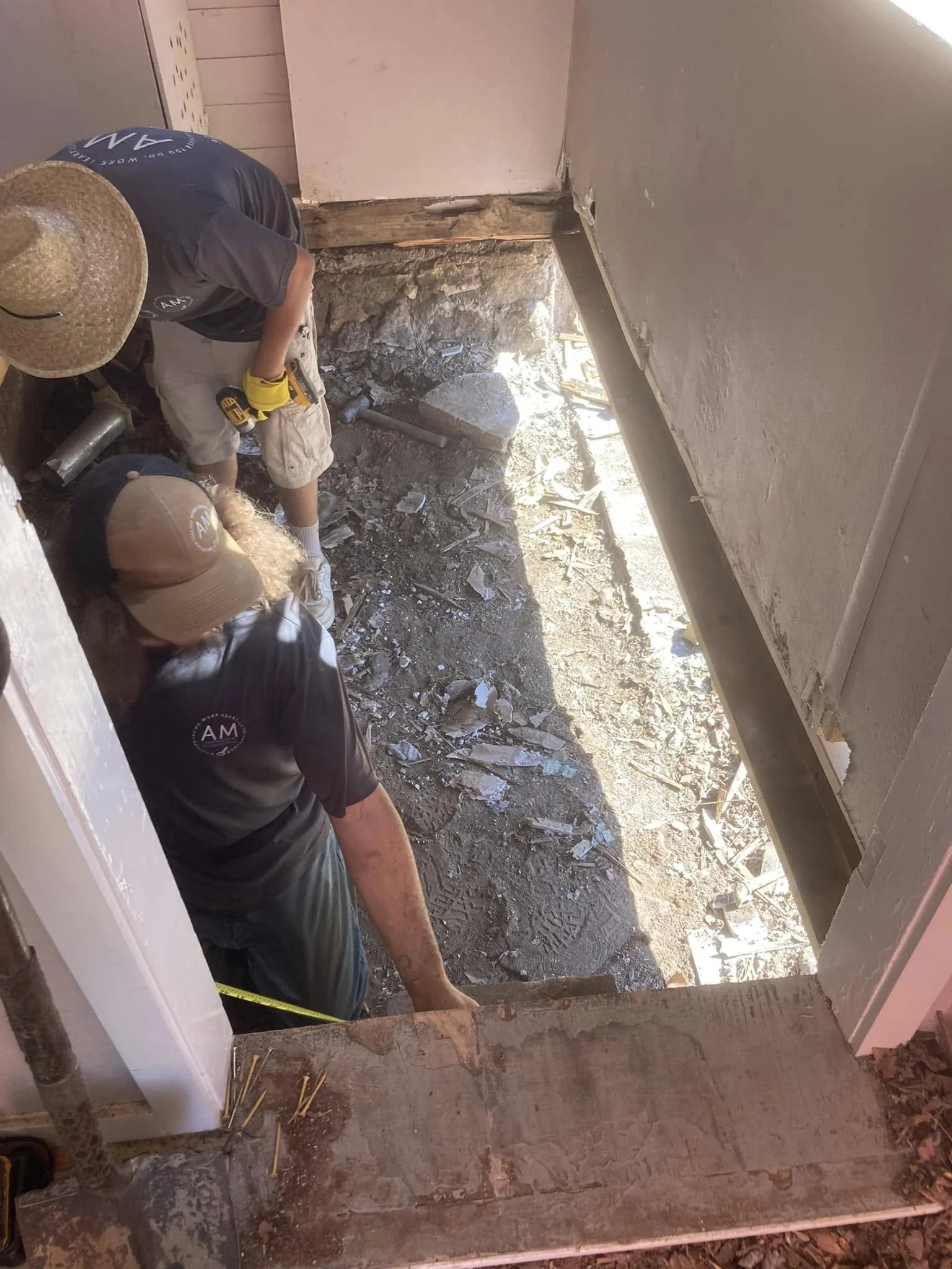 Two workers with hats and work gloves engaged in a doorframe renovation or demolition, exposing the foundation and dirt ground outside.