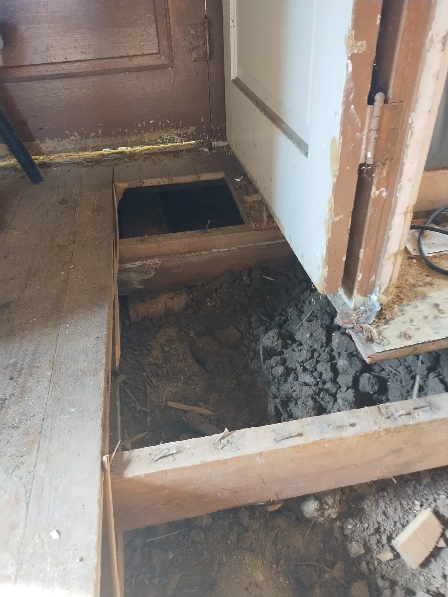 View of a home renovation with a section of the floor removed, revealing soil and supports underneath, with unfinished walls and visible construction debris.