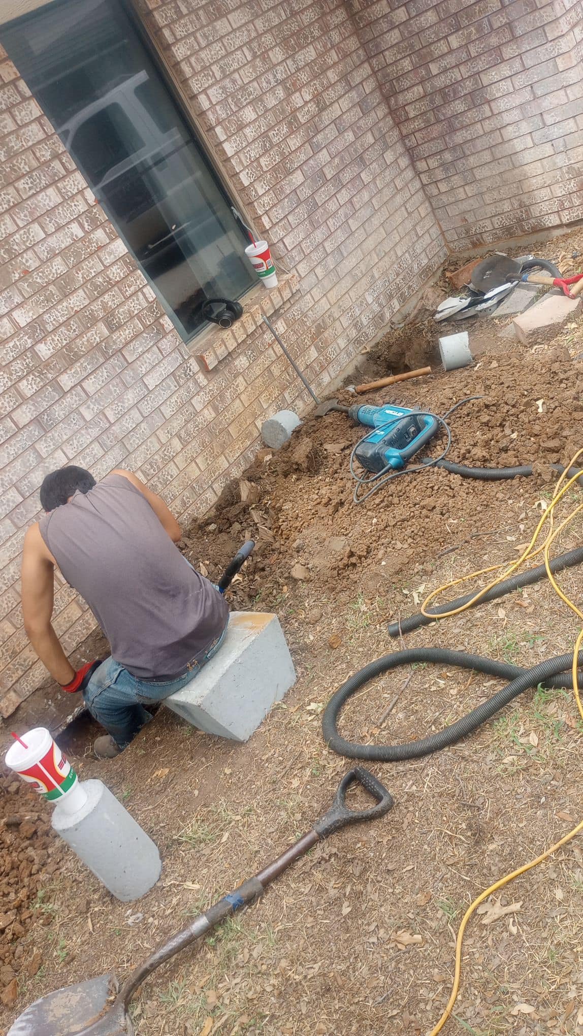 A person working outdoors near a brick house, digging in the dirt with construction tools and equipment, including a power drill, shovel, and hoses.