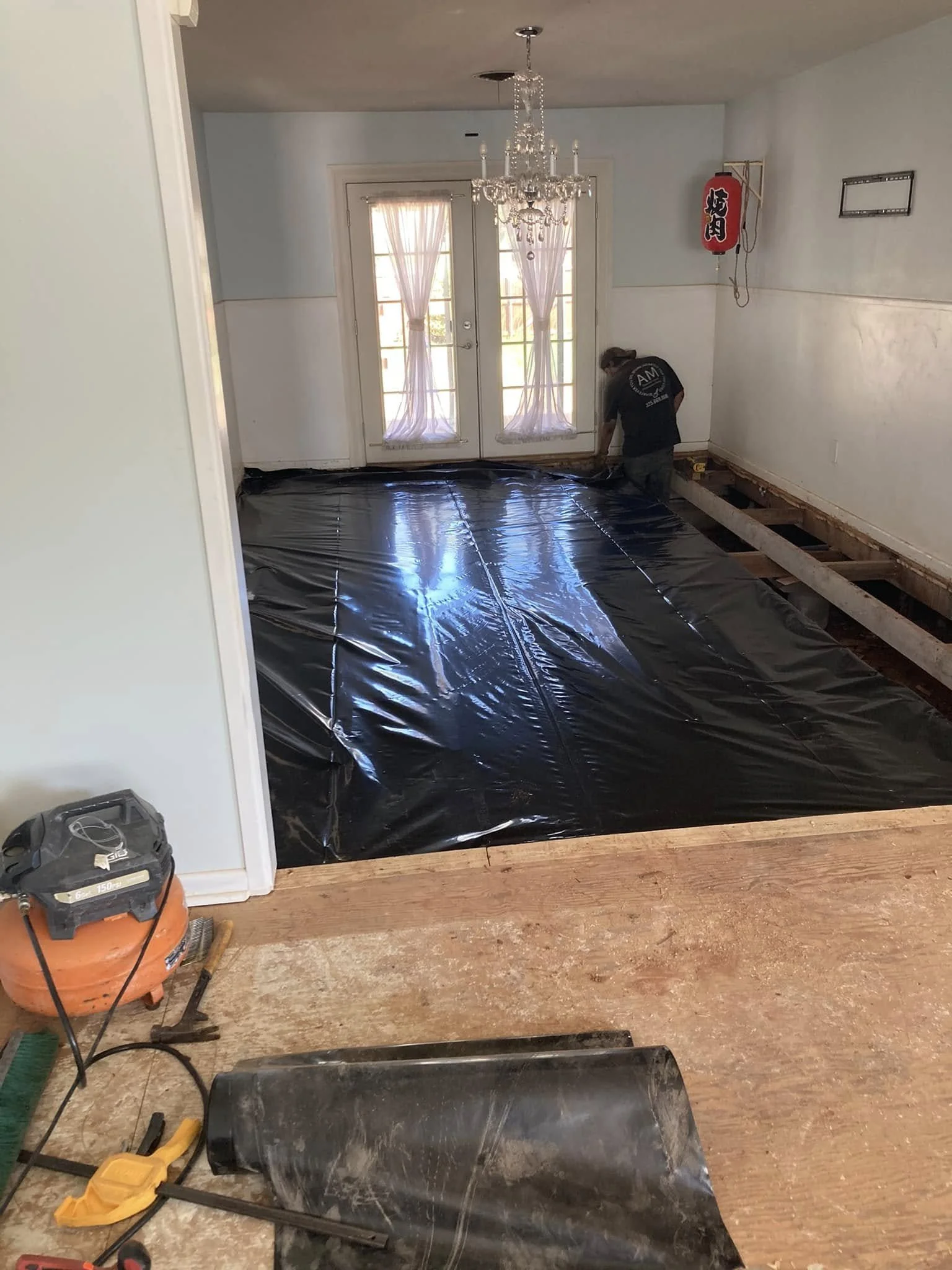 A person working on a home renovation project, installing black plastic sheeting on the floor of a dining room with a chandelier, double doors with pink curtains, and a Japanese lantern on the wall.