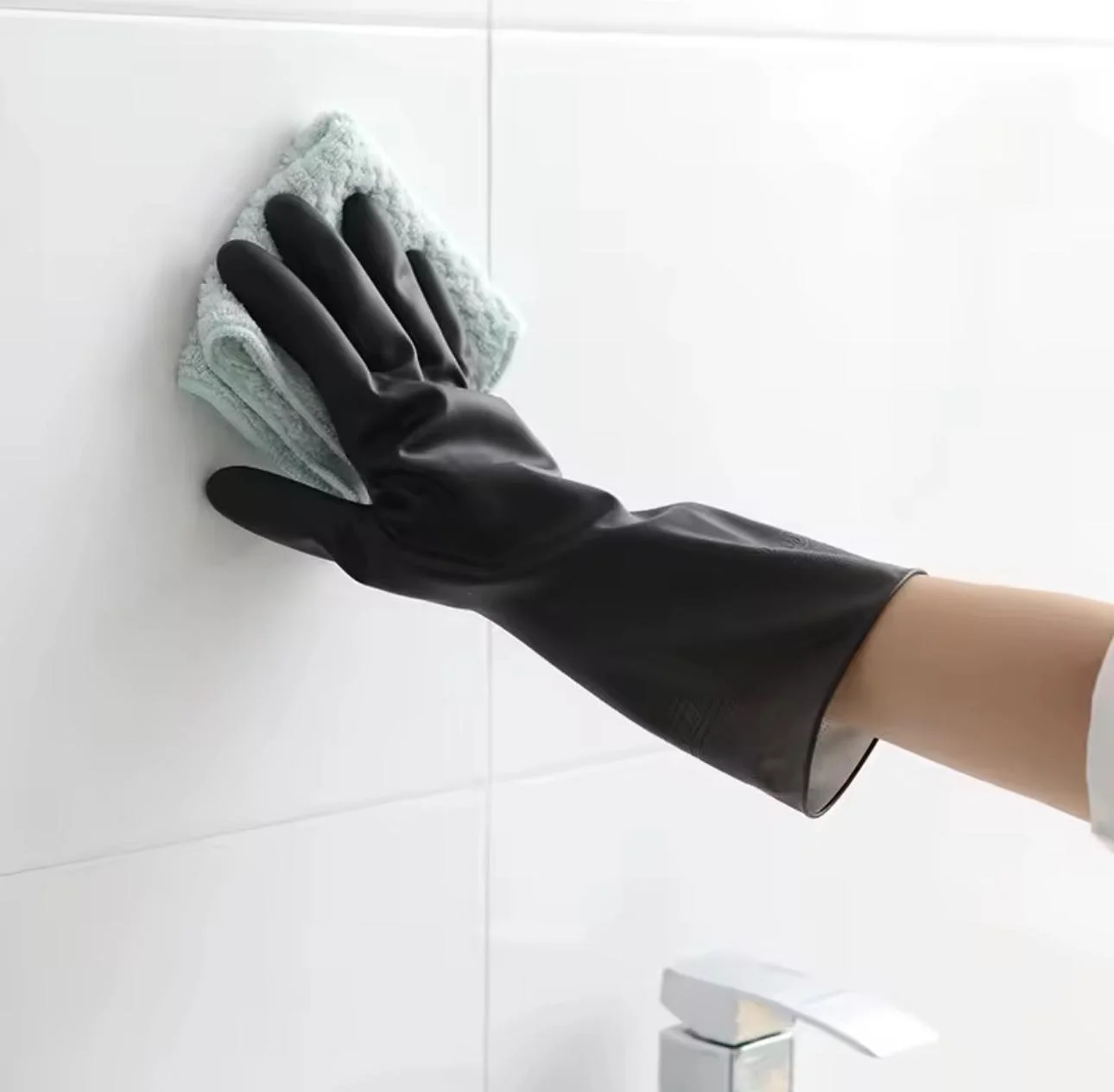 Person wearing a black rubber glove cleaning a white tiled wall with a cleaning cloth.
