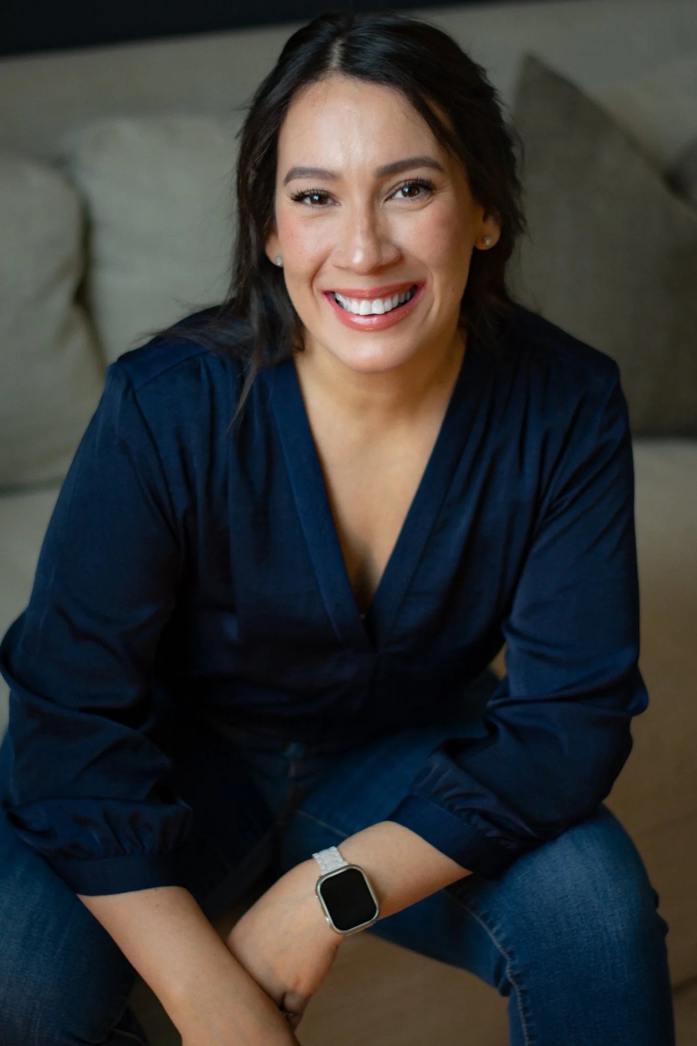 A woman with shoulder-length dark hair, smiling while sitting on a beige couch, wearing a dark blue blouse and jeans, with a smartwatch on her left wrist.