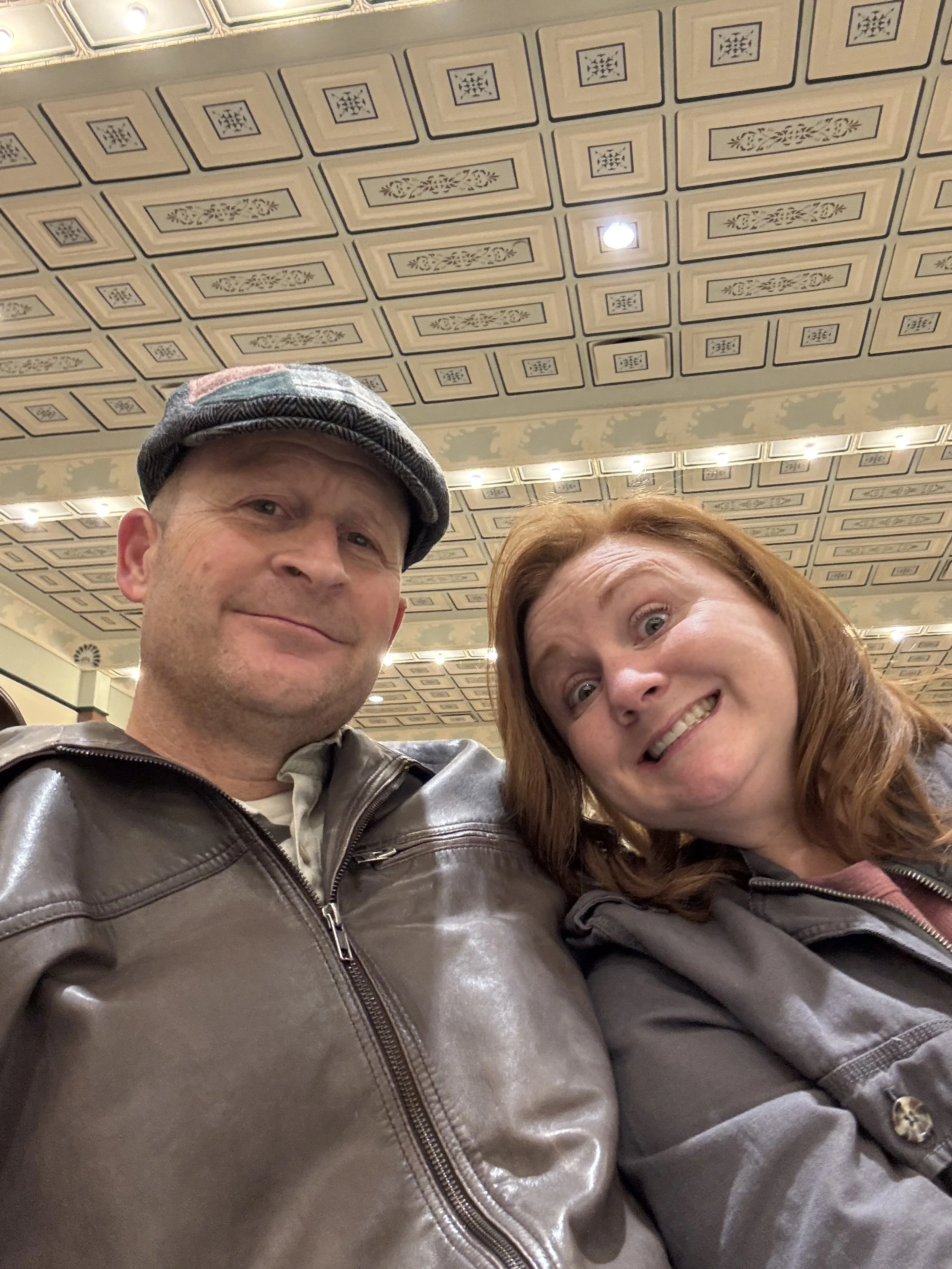 A man and woman taking a selfie indoors. The man is wearing a cap and a brown leather jacket, and the woman has red hair and is wearing a gray jacket. The ceiling has decorative square panels with floral and geometric patterns.