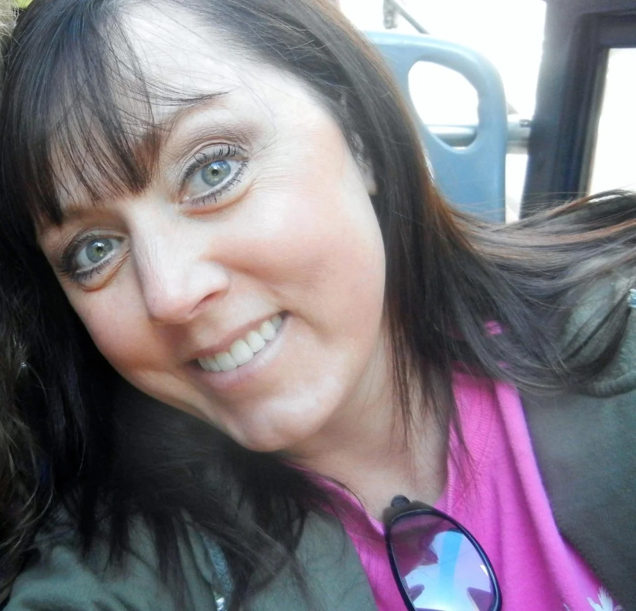 A woman with light-colored eyes and dark brown hair smiling, wearing a pink top and sunglasses hanging from her shirt, sitting on a bus.
