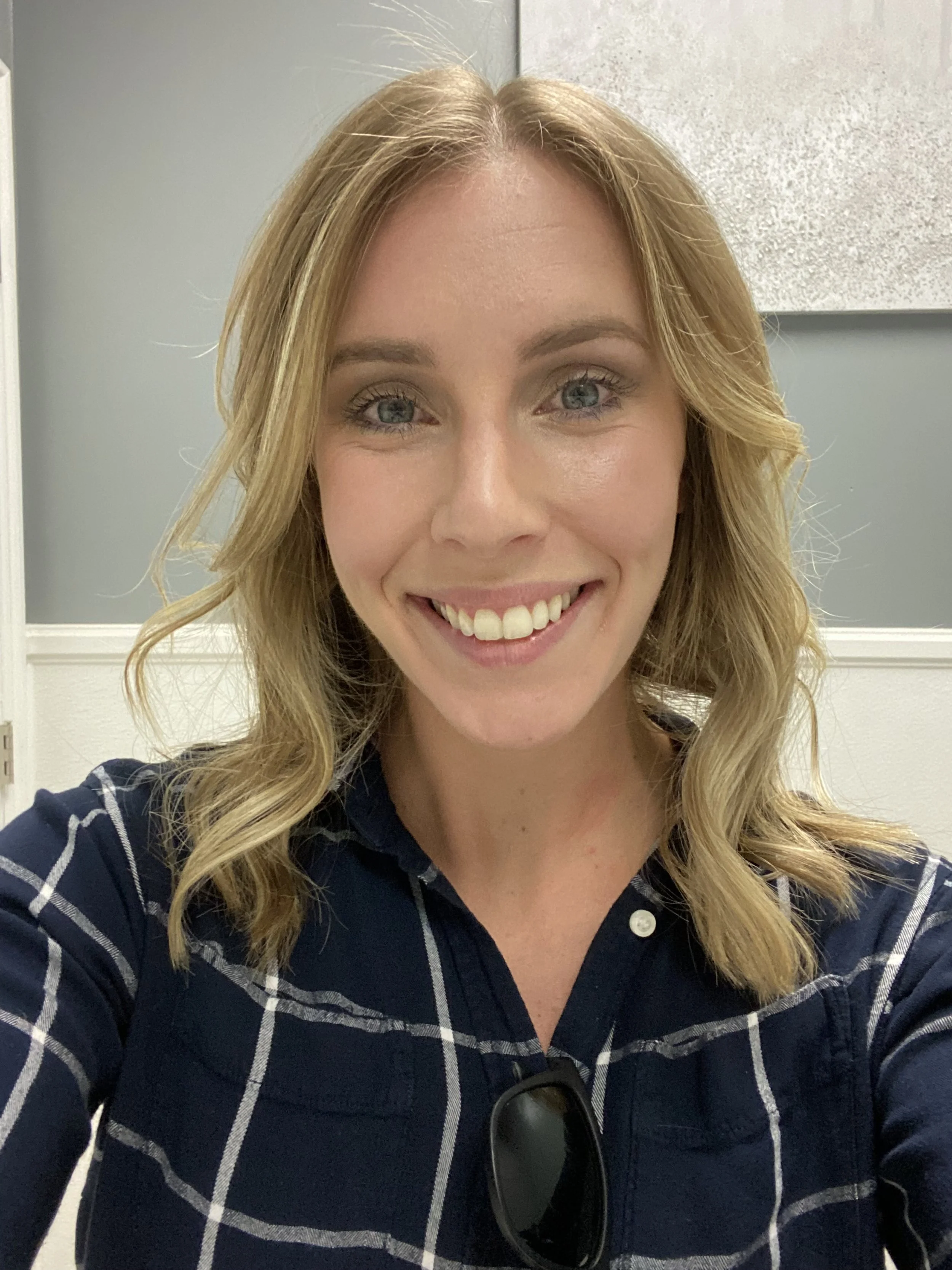 A woman with blonde hair and blue eyes smiling, wearing a navy blue plaid shirt with sunglasses hanging on the shirt, standing in front of a light green and gray wall.