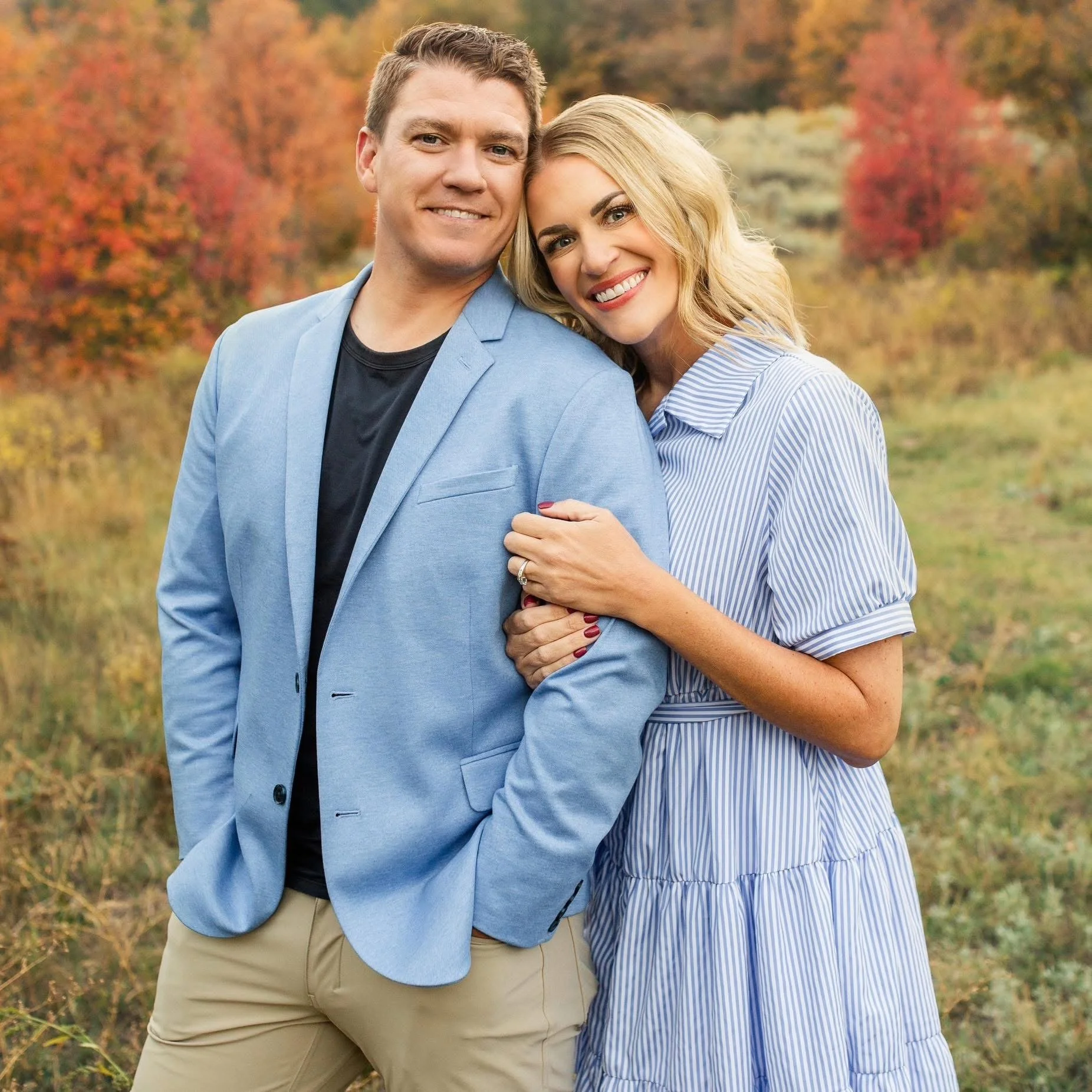 A smiling couple standing close together outdoors during autumn, with colorful fall trees in the background, the woman is holding the man's arm and showing her engagement ring.