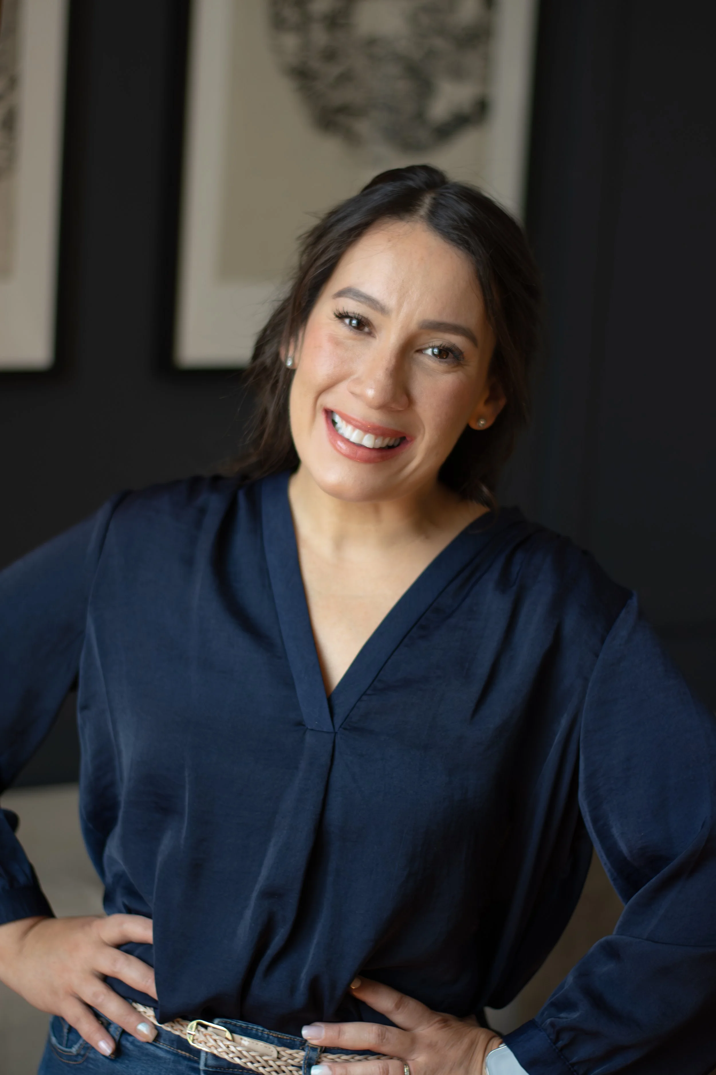 A woman with dark hair smiling and standing with her hands on her hips, wearing a dark blue top, in a room with framed artwork on the wall behind her.