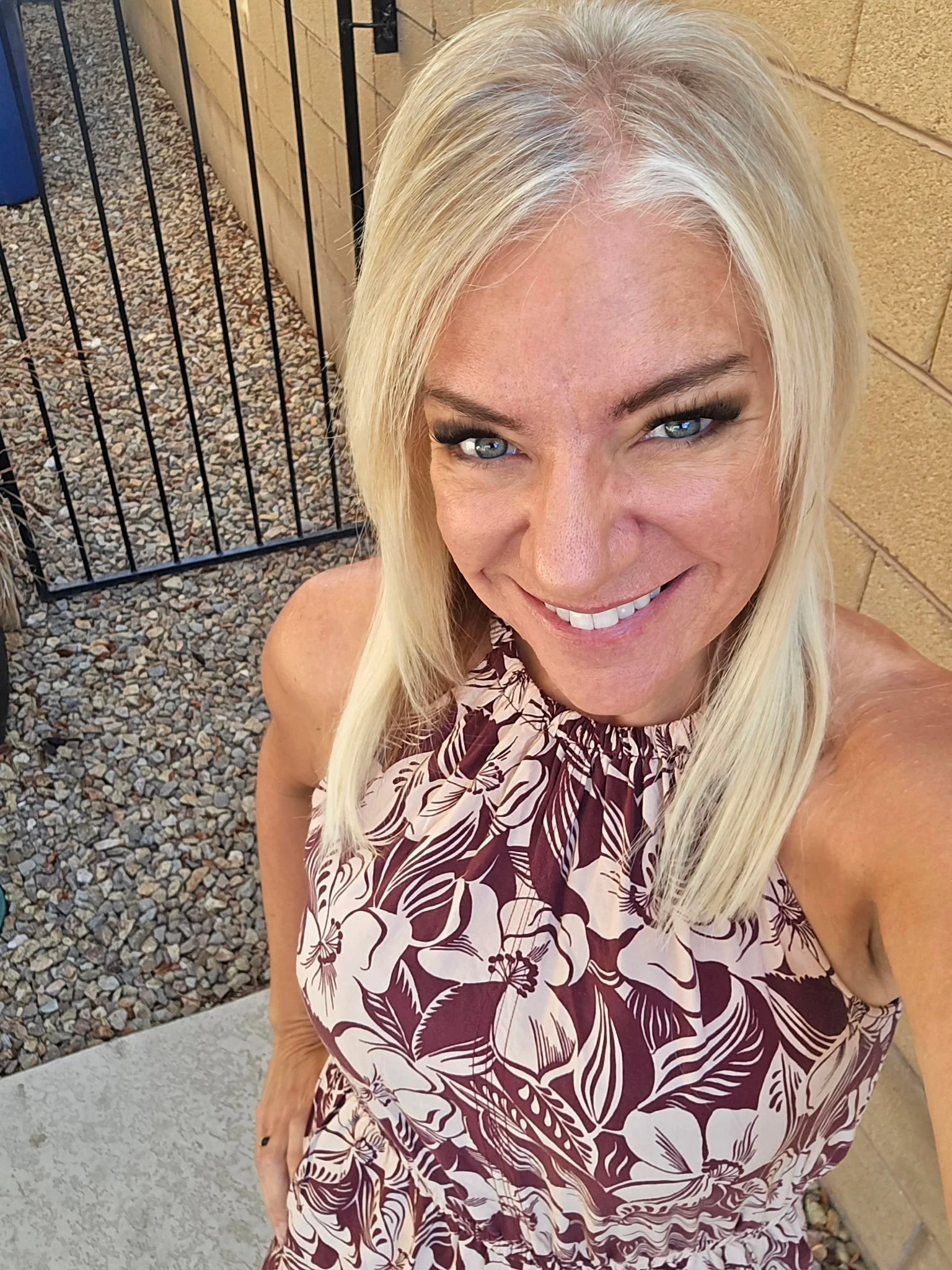 A smiling blonde woman with blue eyes taking a selfie outdoors. She is wearing a sleeveless, floral-patterned dress in shades of purple and white. Behind her, there is a concrete pathway, a gravel area, a black metal fence, and a beige brick wall.