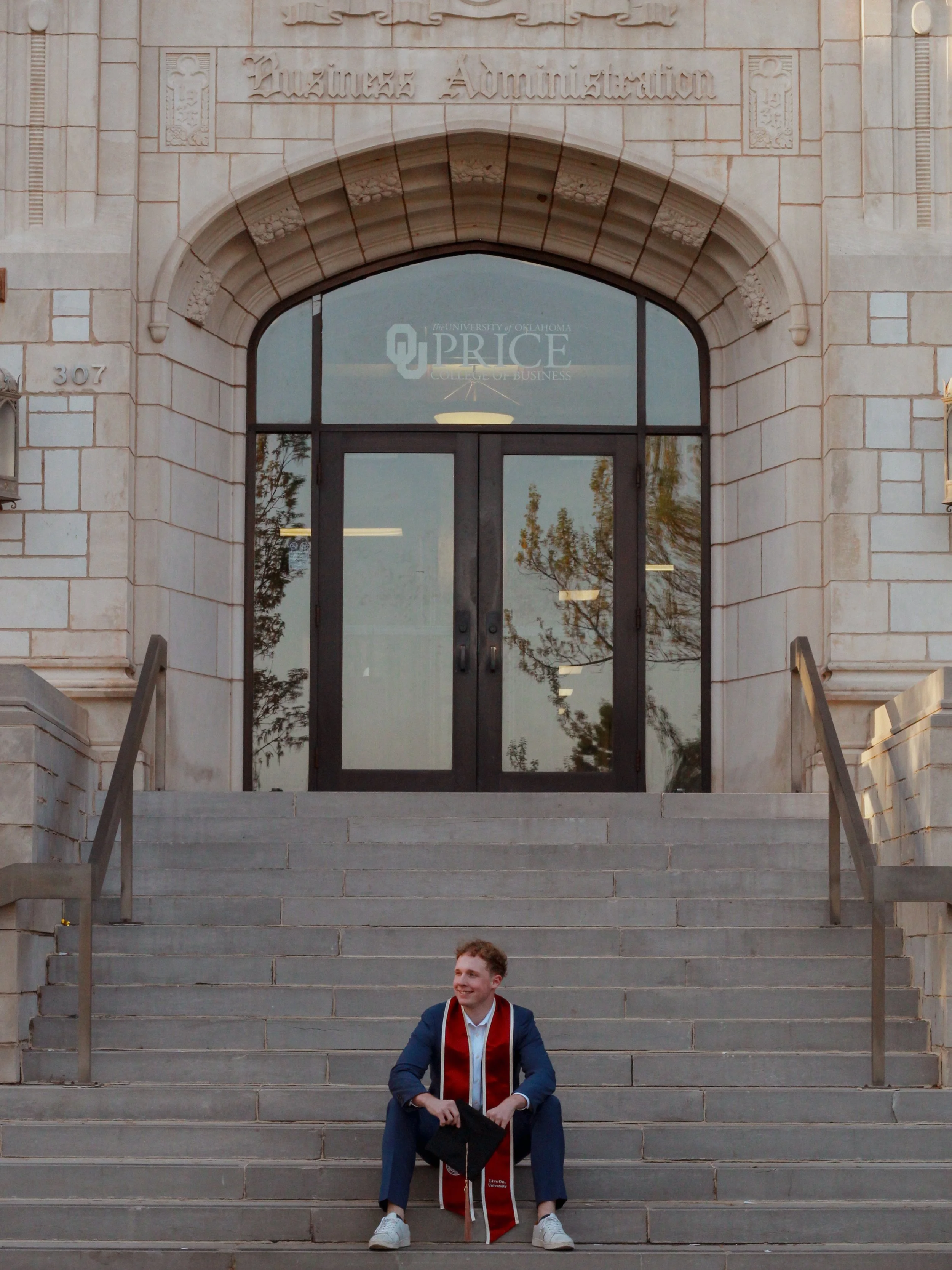 A male OU College Grad sitting on the stairs of Price College during sunset