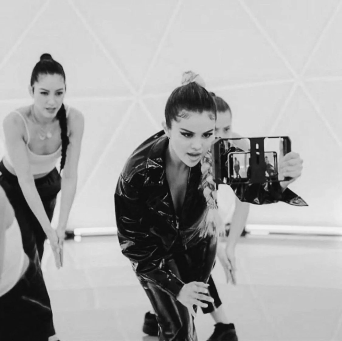 A black-and-white photo of a woman in a stylish outfit holding a camera, capturing herself and others in a studio setting.