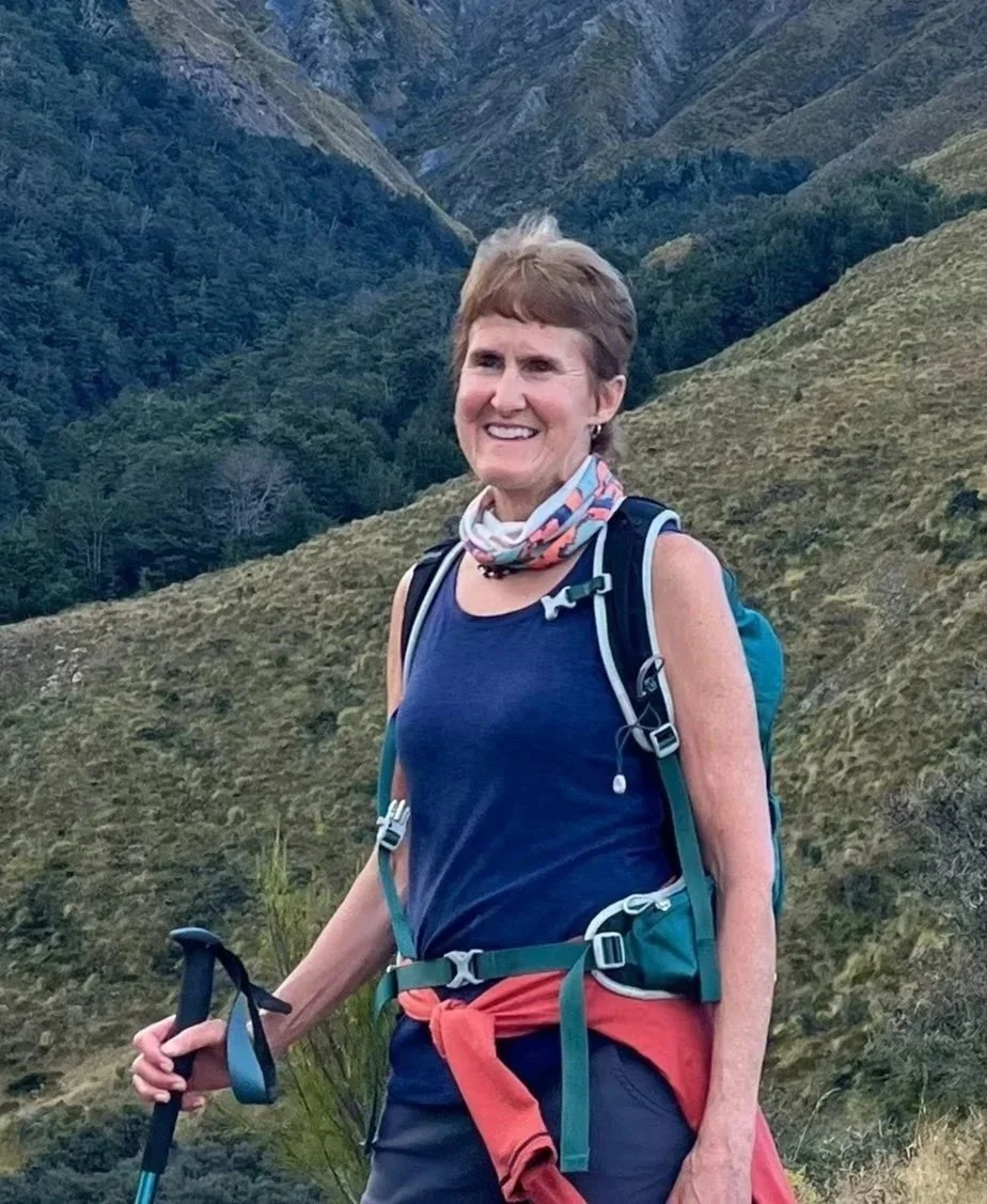 A woman with short brown hair smiling, carrying a backpack, and holding trekking poles, standing outdoors in a mountainous area.