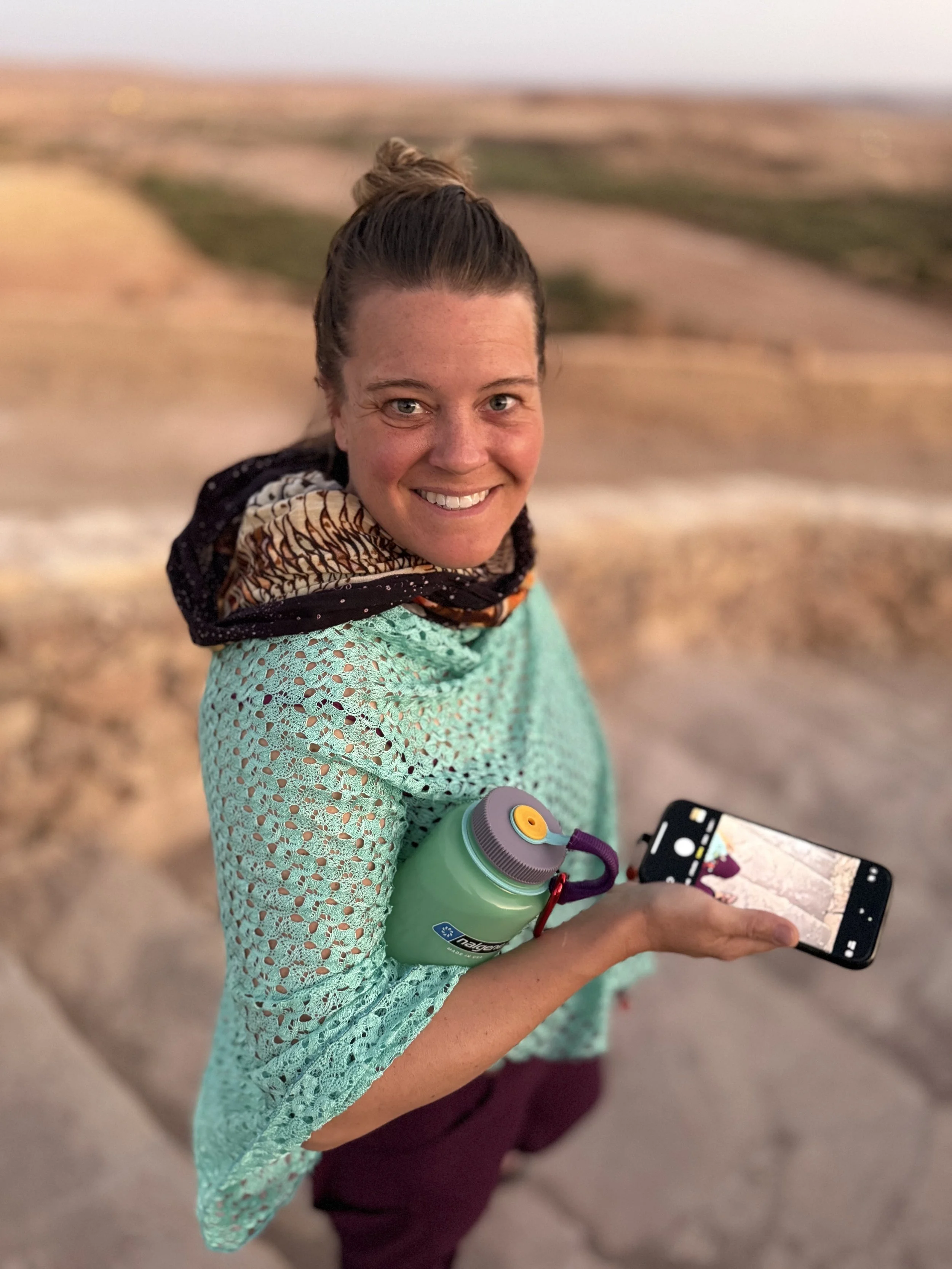 Woman smiling outdoors, holding a smartphone and a green water bottle, with a scenic landscape background.