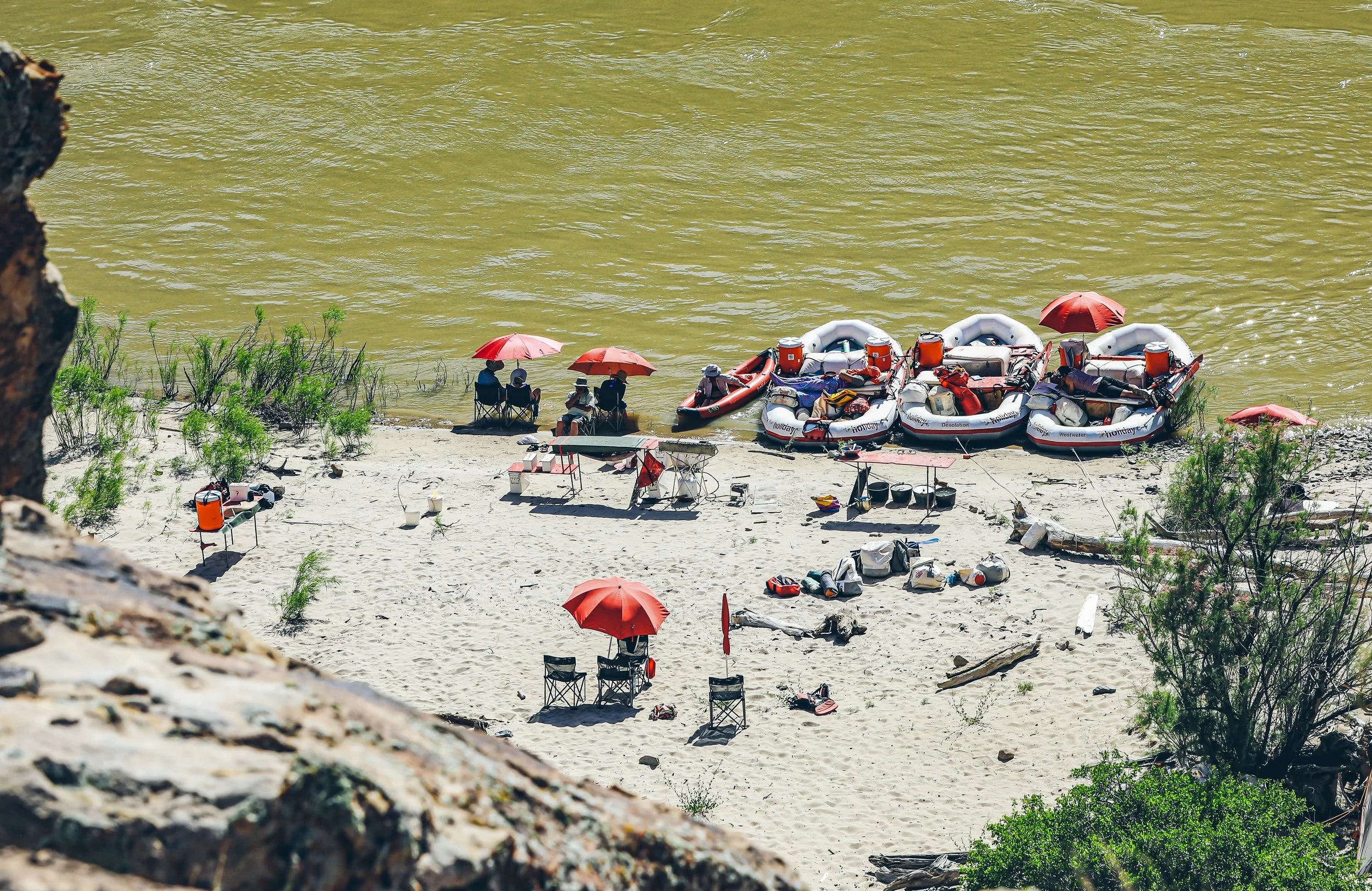 A beach scene with people sitting under red umbrellas, some in boats, and various supplies scattered on the sand, seen from a higher vantage point.