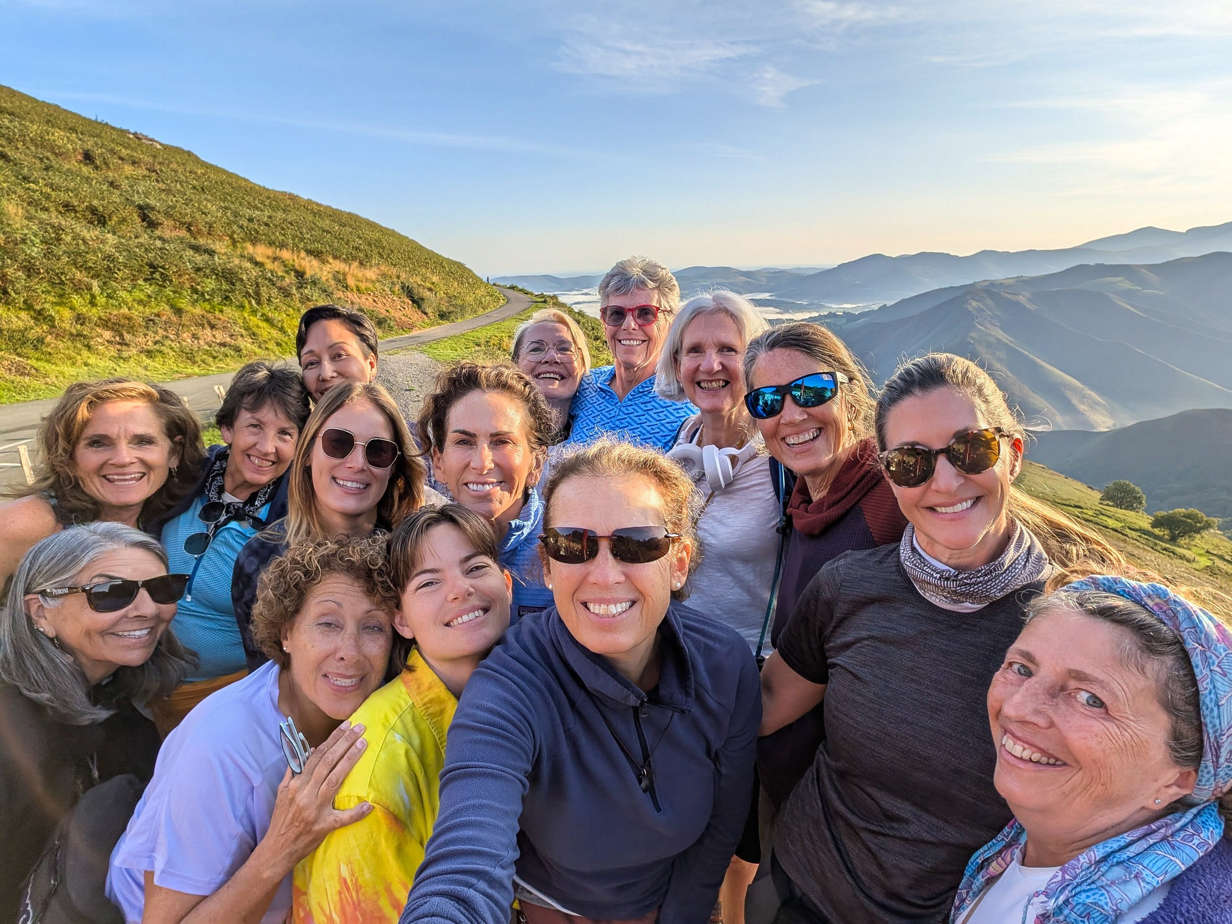 Group of 16 women smiling together outdoors on a mountain trail with rolling hills and a valley in the background, during sunset.