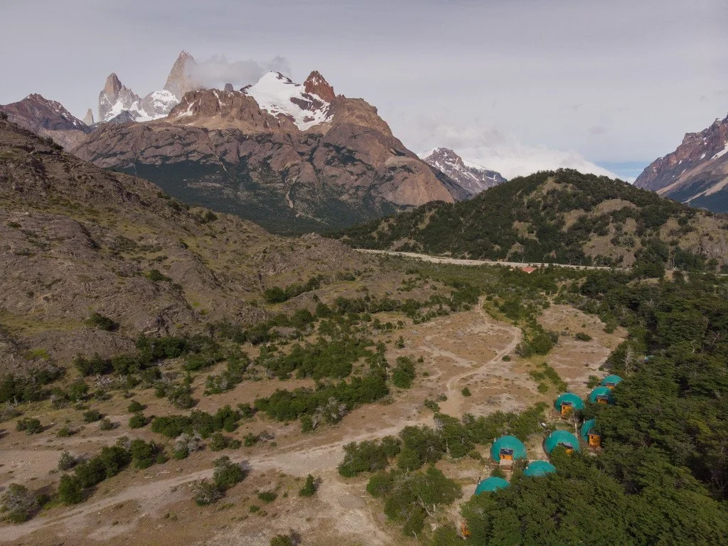 A mountain landscape with snow-capped peaks and rugged terrain, featuring a small cluster of teal tents on the lower right side.
