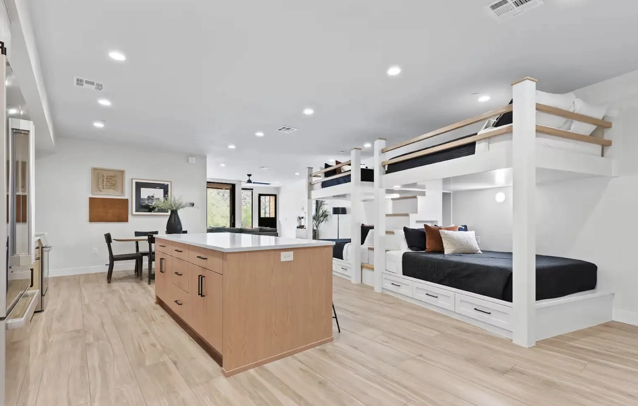 Open-concept living space with a kitchen island, dining area, and built-in bunk beds in the background, with large windows and modern decor.