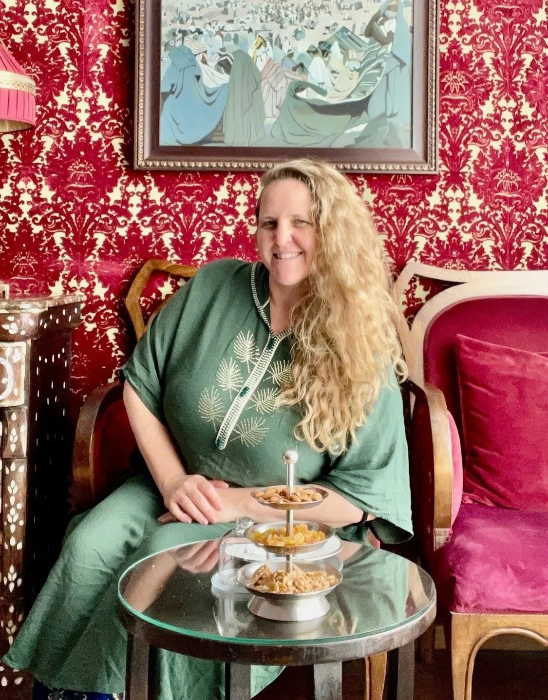 A woman with long, curly blonde hair smiling and sitting at a table with a three-tiered tray of snacks in a room with red damask wallpaper and framed artwork behind her.