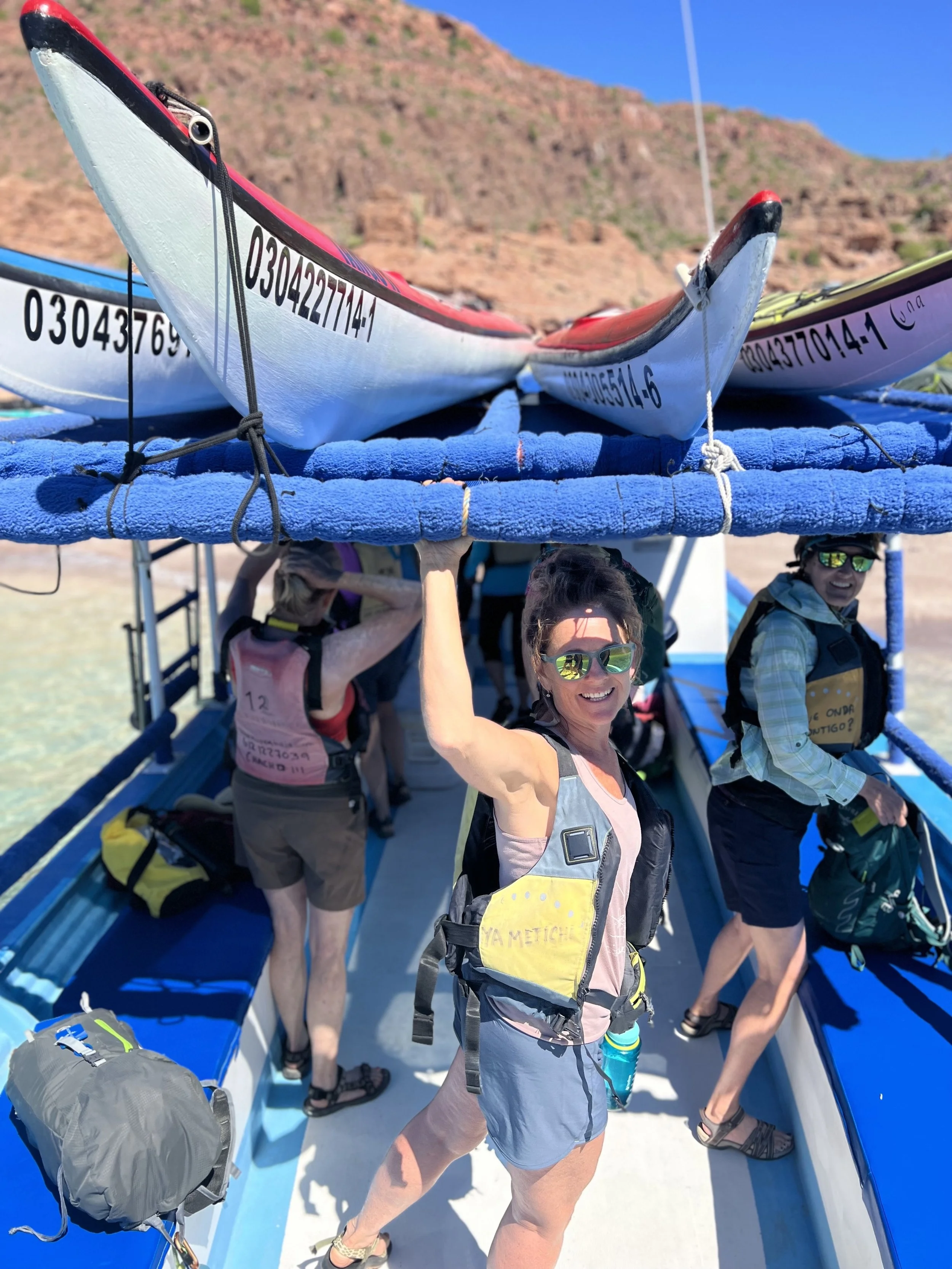 Group of women sitting on a boat with hills in the background, some wearing life jackets, sunglasses, and hats, on a sunny day.