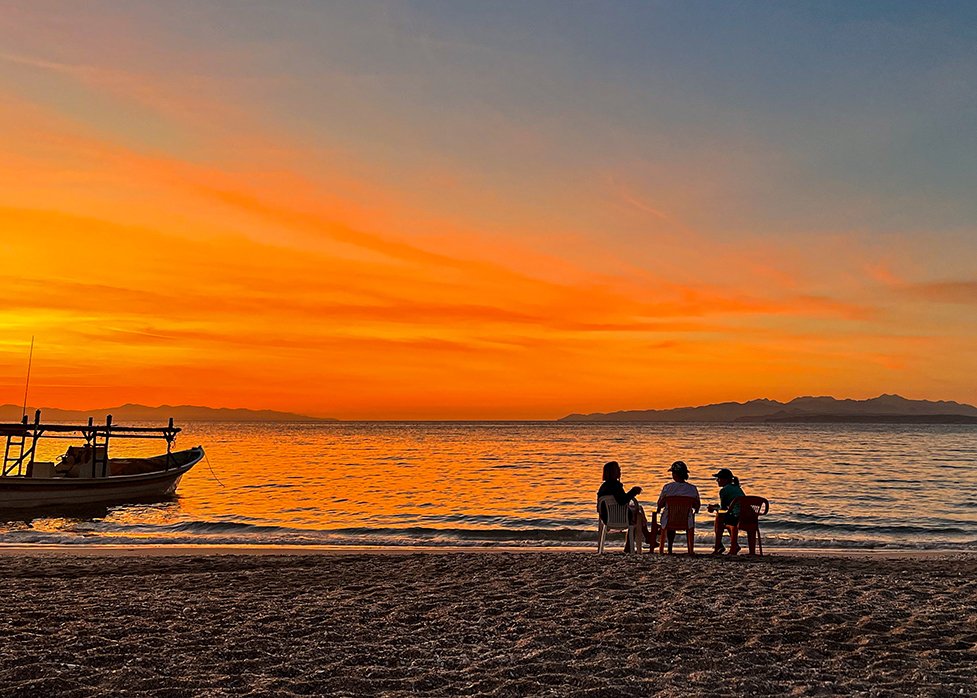 Three people sitting on chairs on a beach at sunset, with a boat on the water to the left and an island or landmass in the distance.