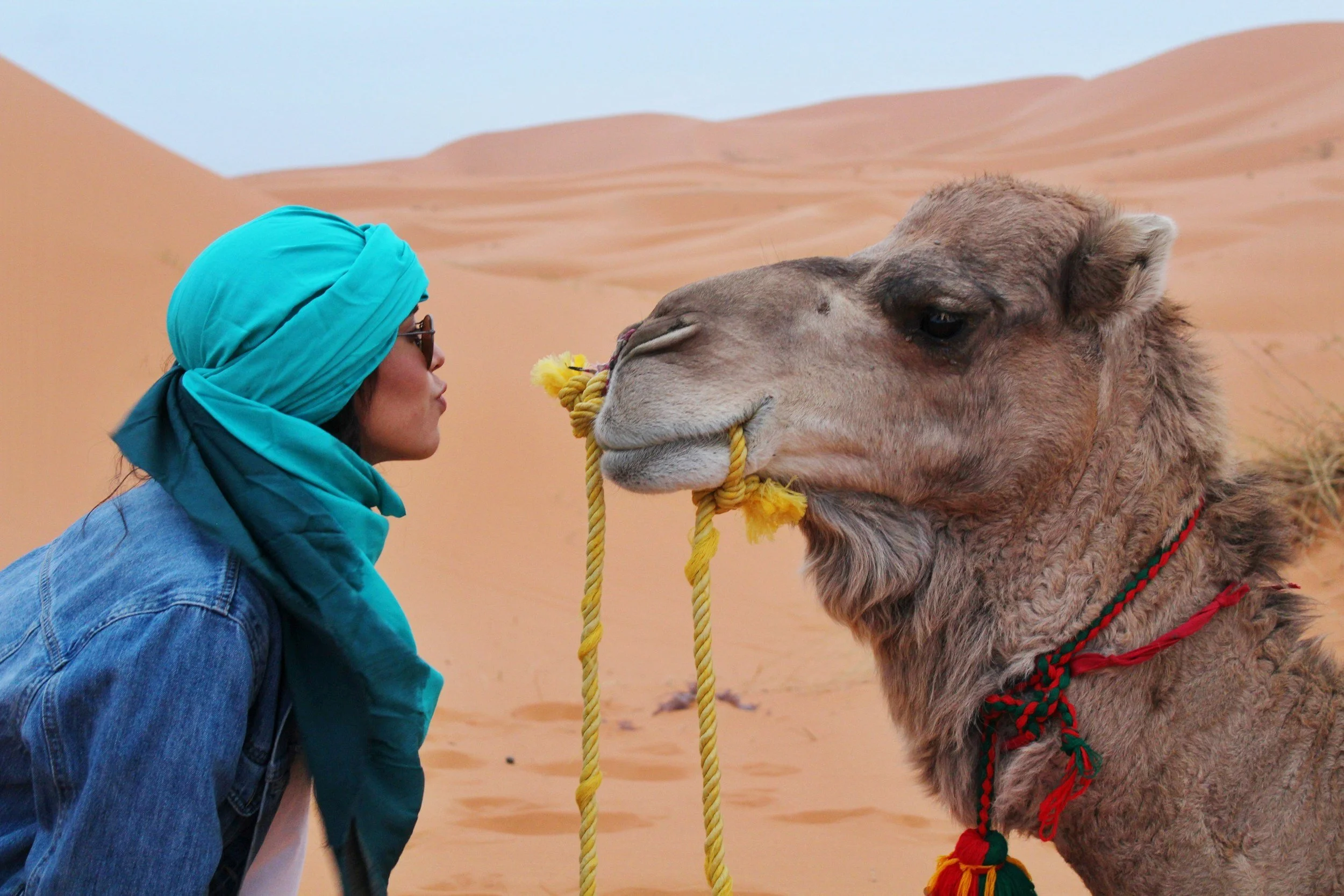 A woman wearing a blue headscarf and sunglasses is nose-to-nose with a camel in a desert landscape with sand dunes.