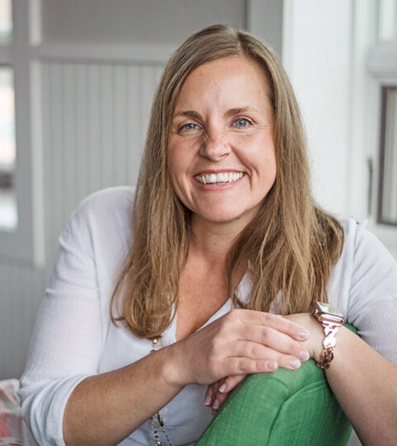 A woman with long blonde hair and blue eyes smiling, sitting in a well-lit room with light-colored walls, wearing a white shirt and a watch on her left wrist.