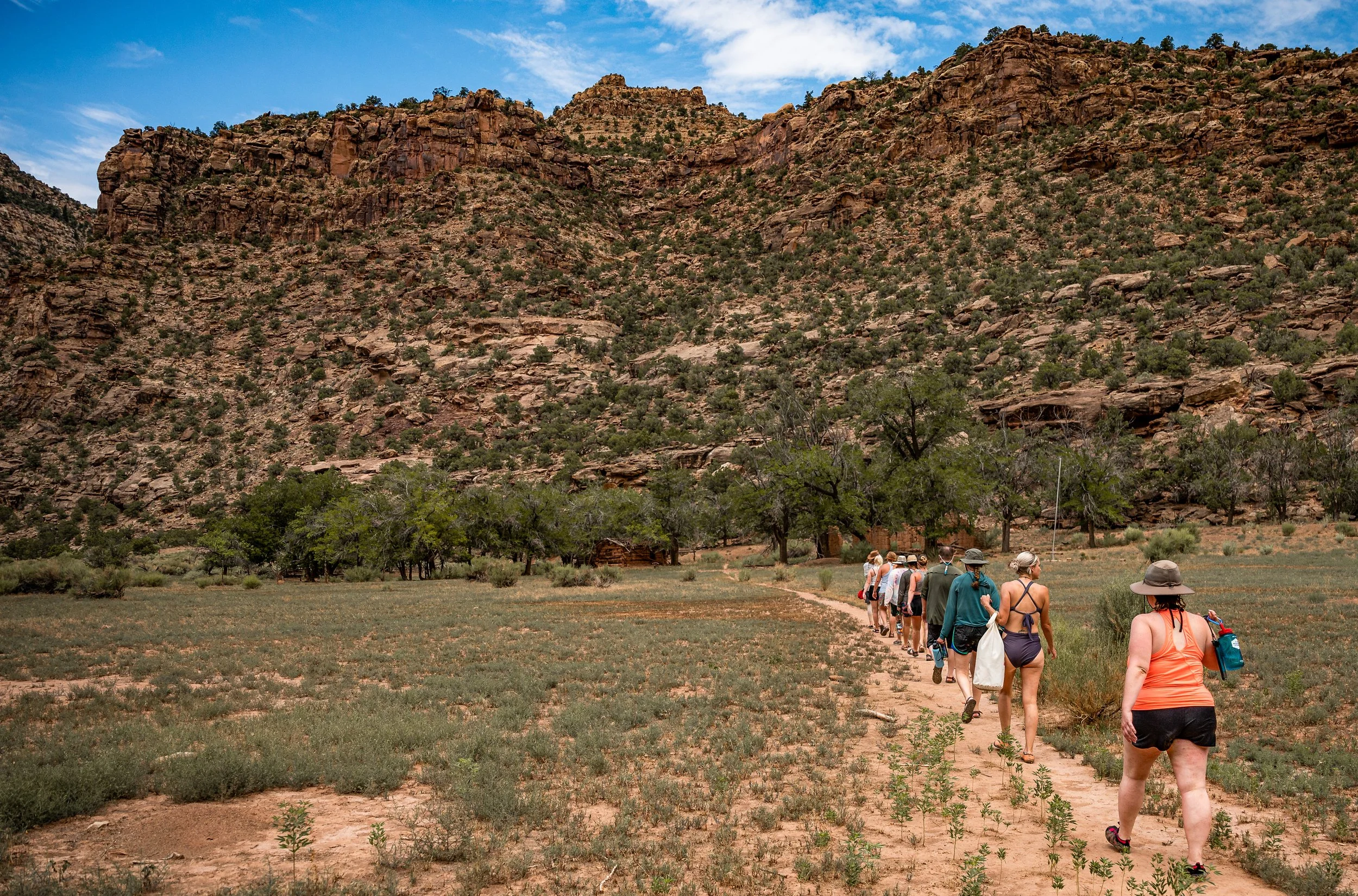 A group of hikers walking single file on a dirt trail through a semi-arid landscape with sparse vegetation, trees, and tall rocky hills under a partly cloudy sky.