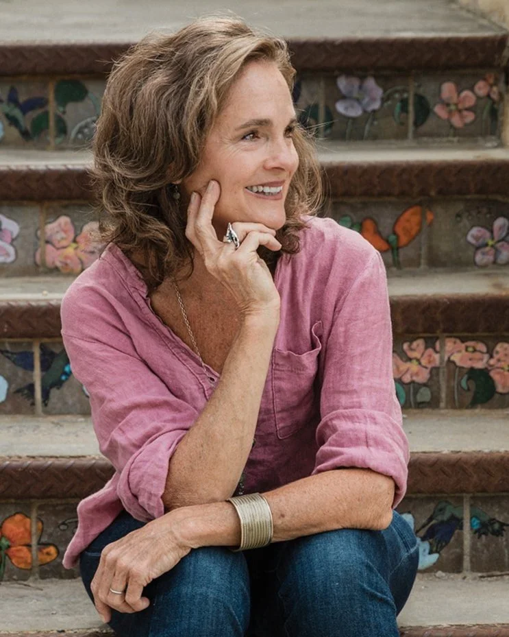 A woman with curly brown hair sitting on decorated steps with floral and butterfly designs, smiling and looking to the side, wearing a pink shirt, jeans, and jewelry.