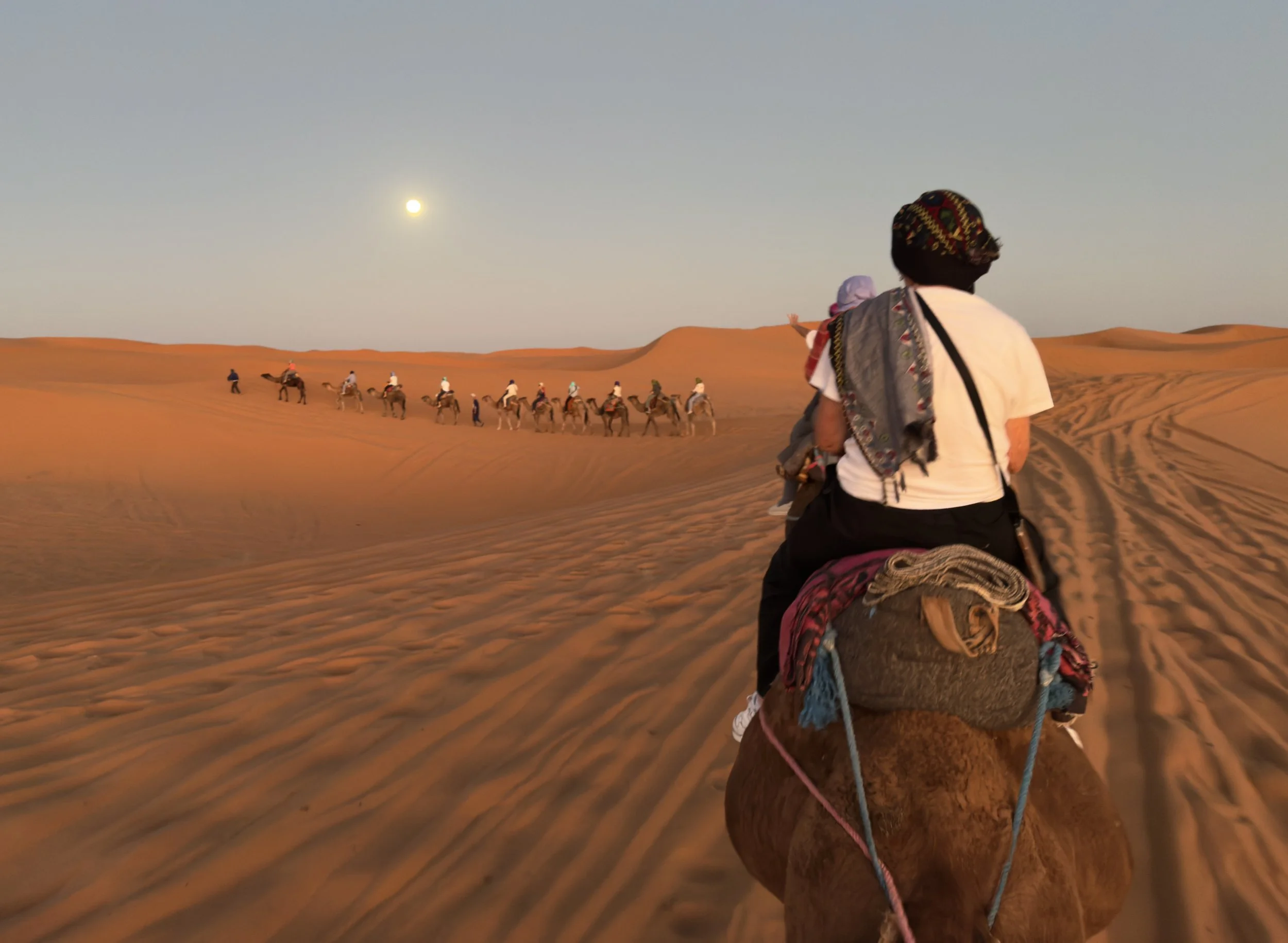 People riding camels in a desert at sunset with a full moon in the sky.