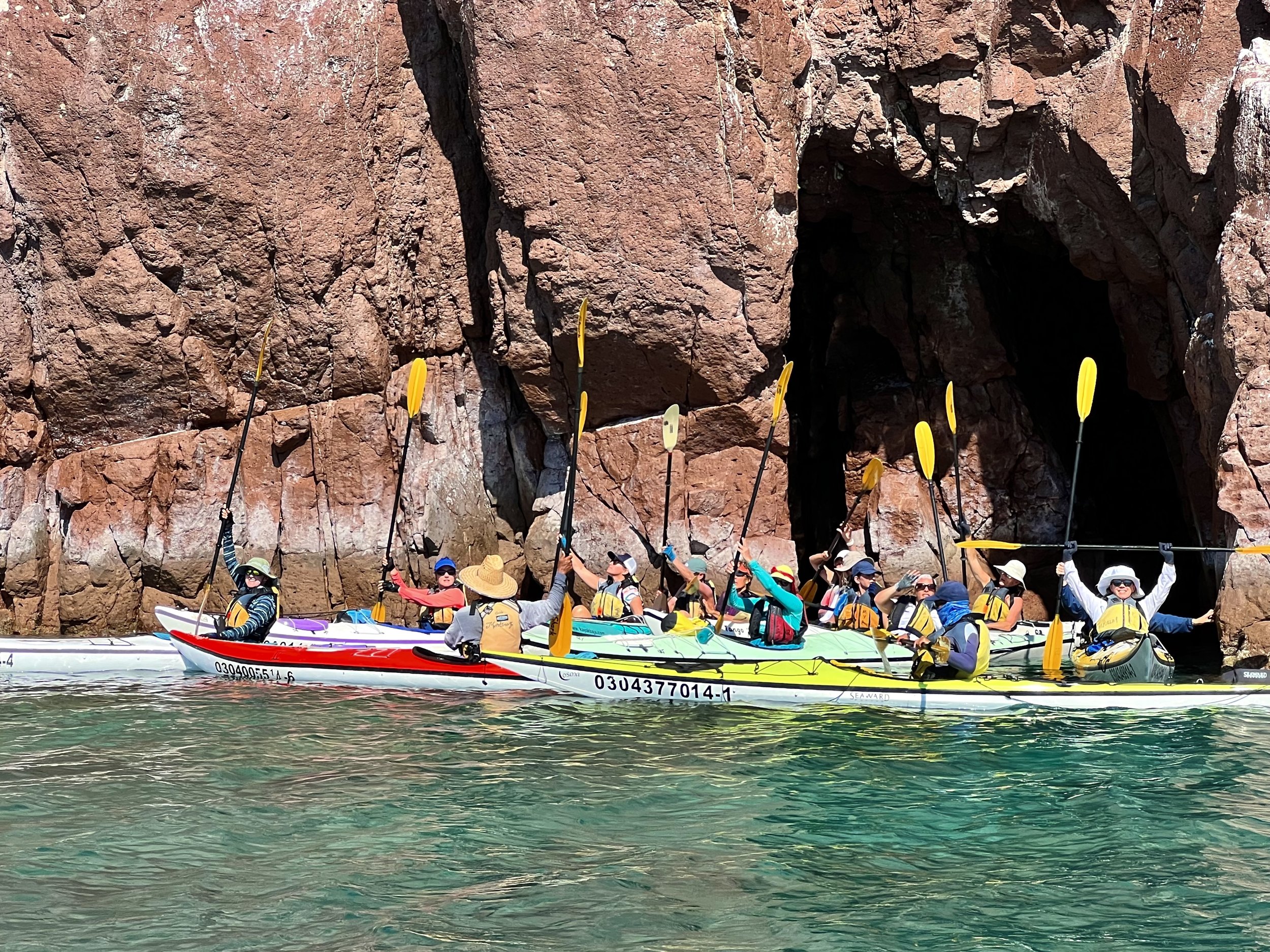 A group of people in kayaks paddling near rocky cliffs, some wearing hats and sunglasses, with a cave opening in the background.