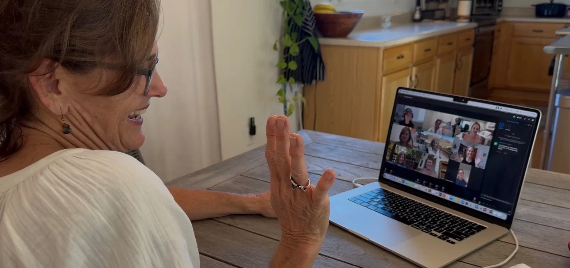 A woman smiling and waving at her laptop screen during a video call. She is sitting at a wooden table in a kitchen with wooden cabinets and a potted plant in the background.