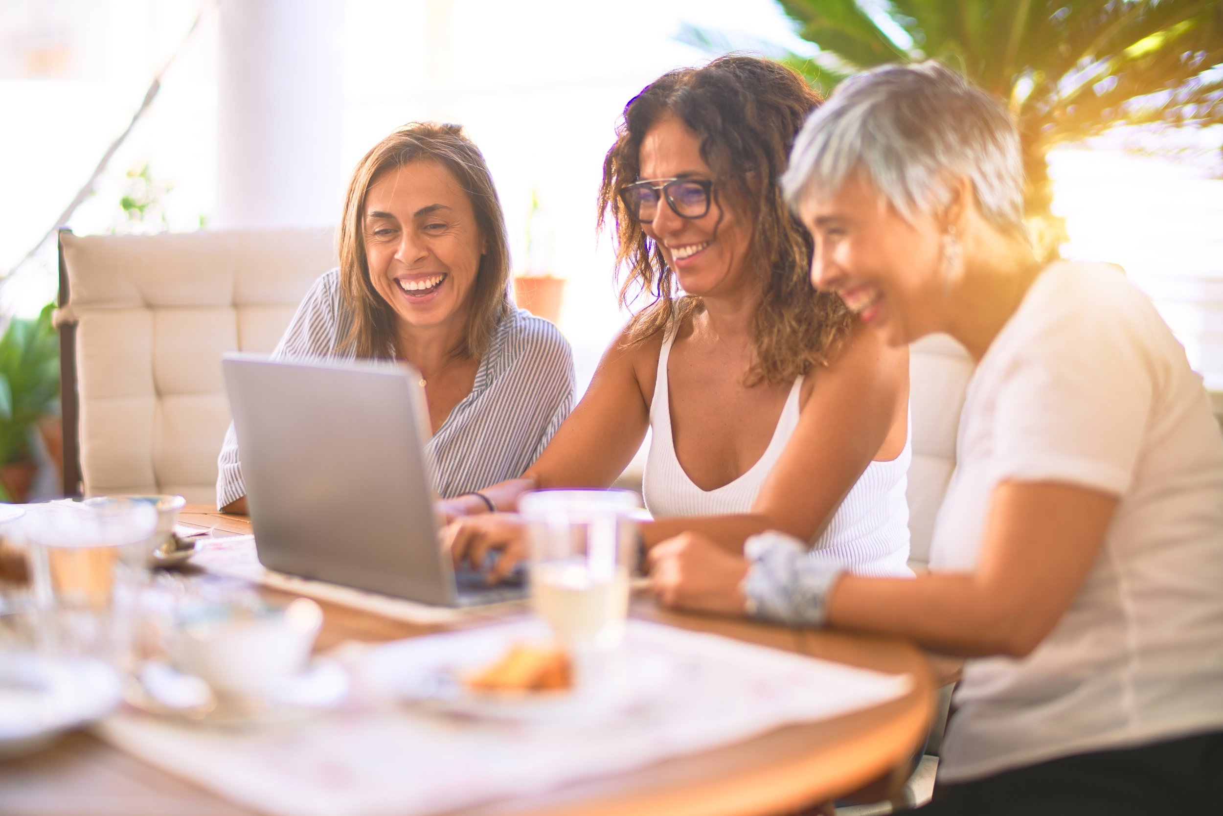 Three women sitting at a table, looking at a laptop, smiling and enjoying a conversation in a bright, sunny room.