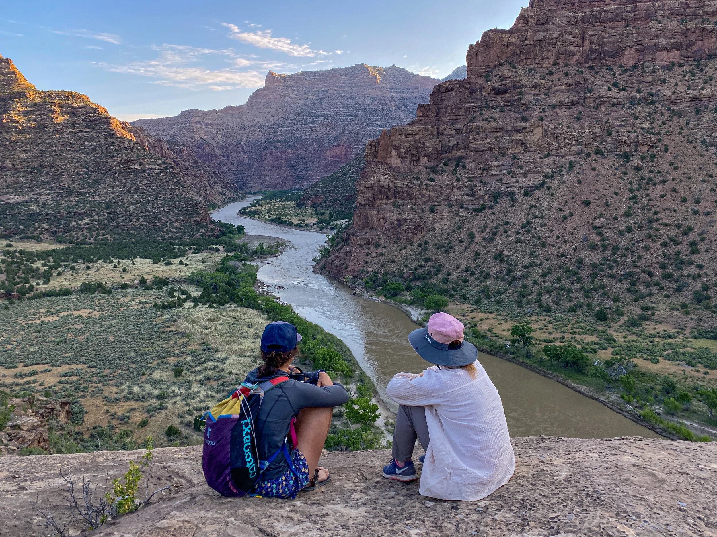Two hikers sitting on a cliff overlooking a river flowing through a canyon with steep cliffs and a partly cloudy sky.