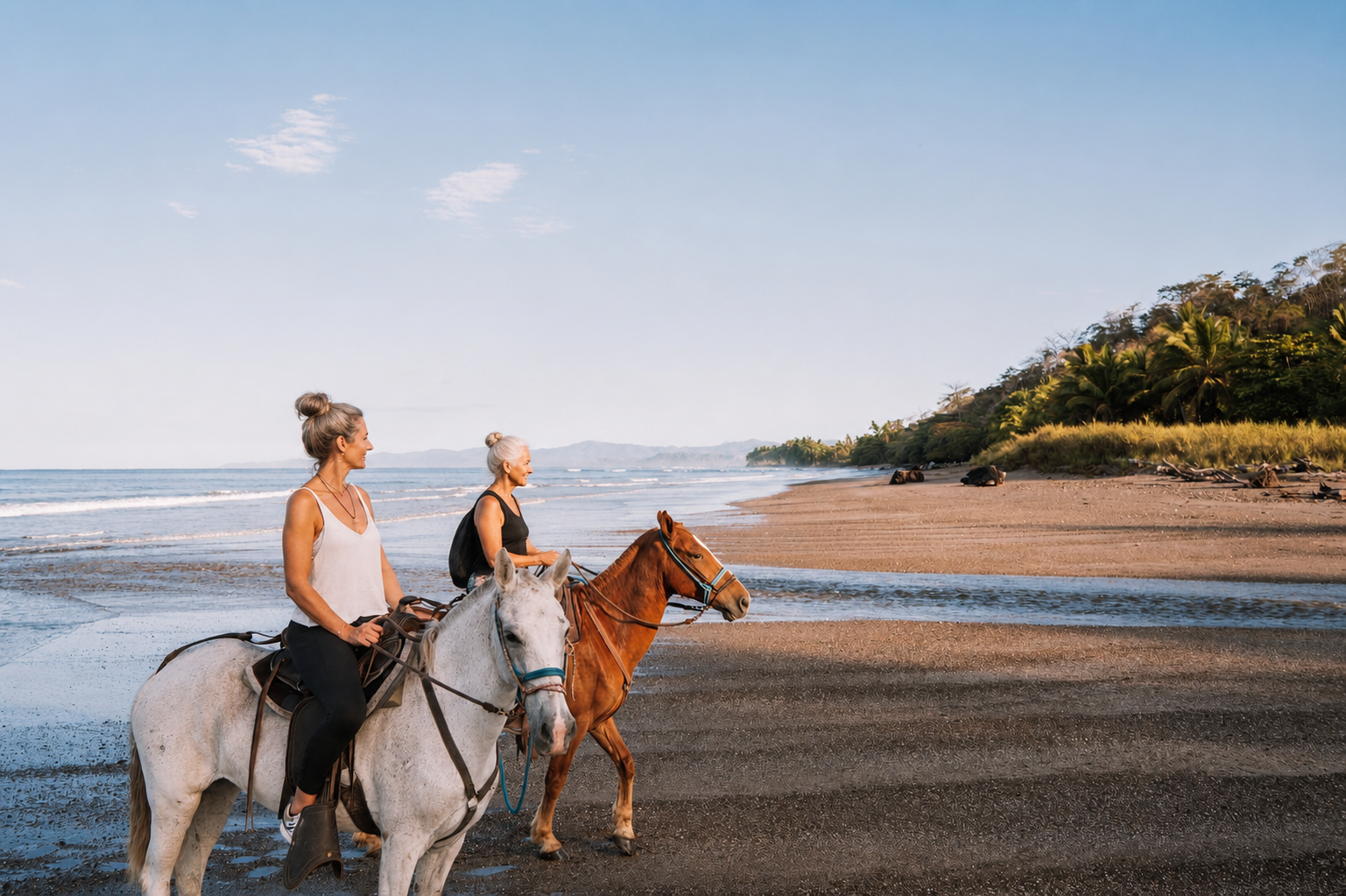 Two women riding horses along a beach with the ocean on their left and a green hillside on their right under a blue sky.