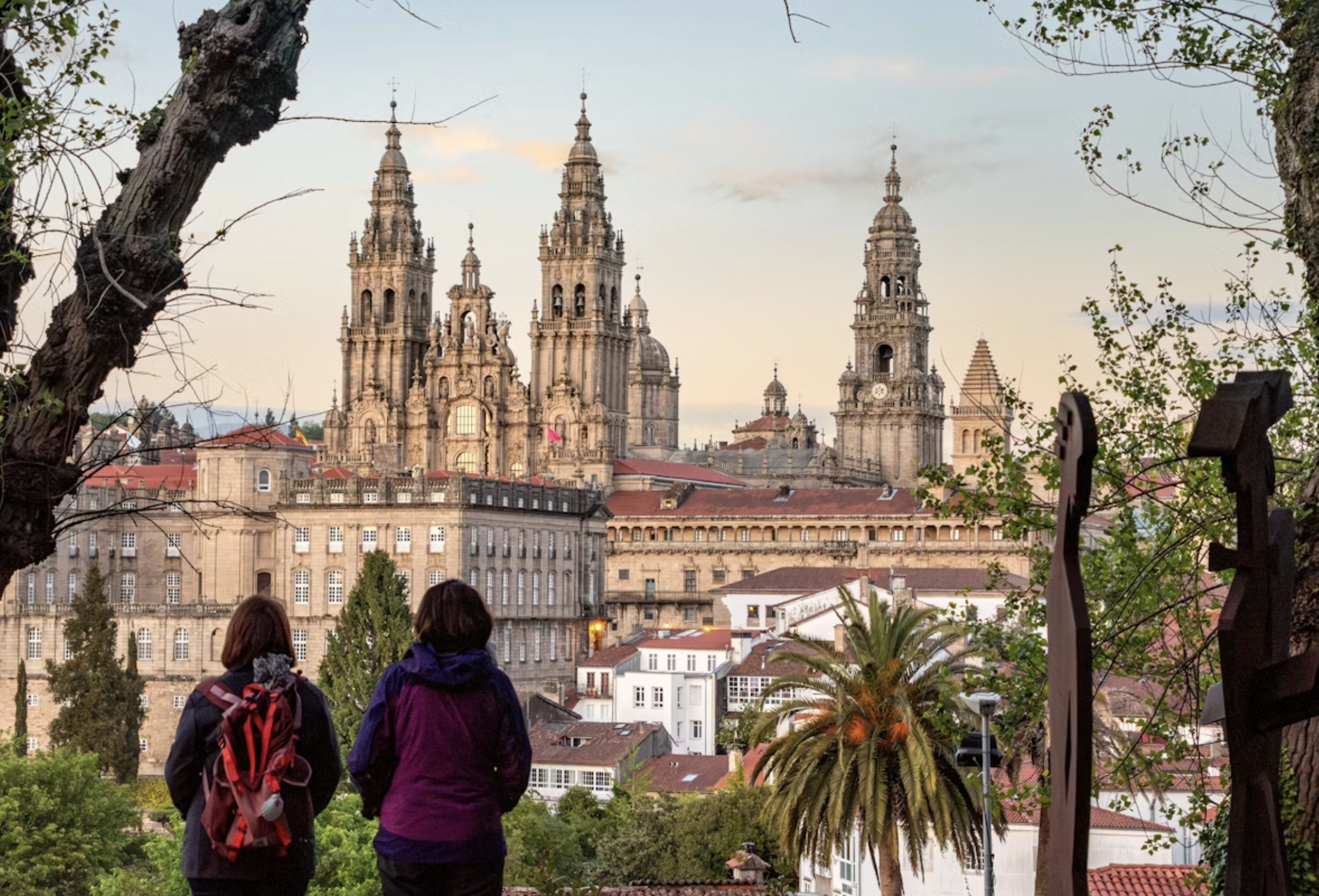 Two people with backpacks standing and looking at a city skyline featuring large historic cathedral towers, with trees and buildings in the foreground.