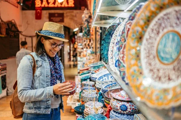 A woman shopping for decorative plates at a store. She is wearing a straw hat, glasses, a denim jacket, and a blue scarf.