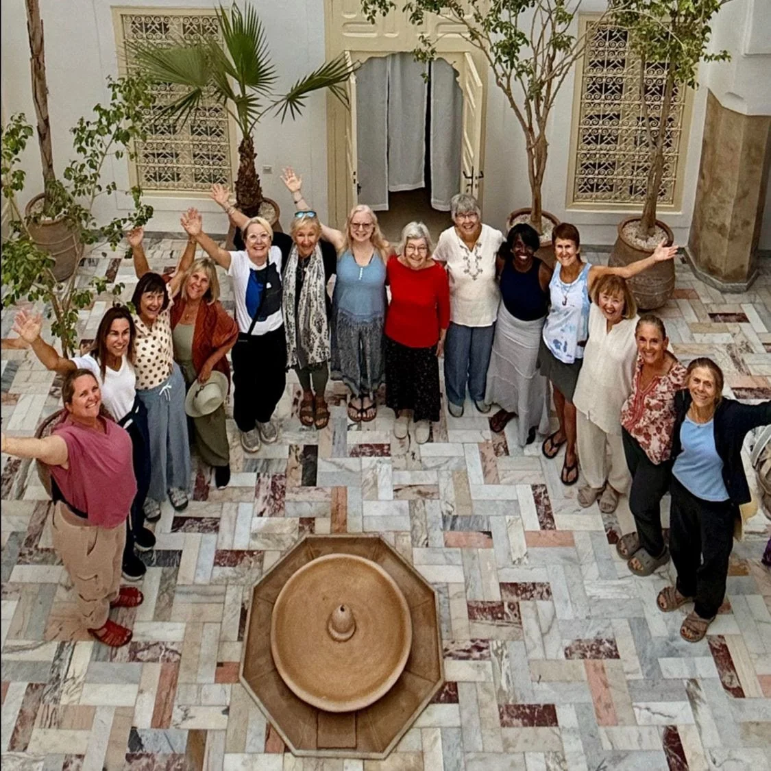 Group of people standing in a circle, smiling and waving, in a decorated indoor courtyard with potted plants and tiled floor.