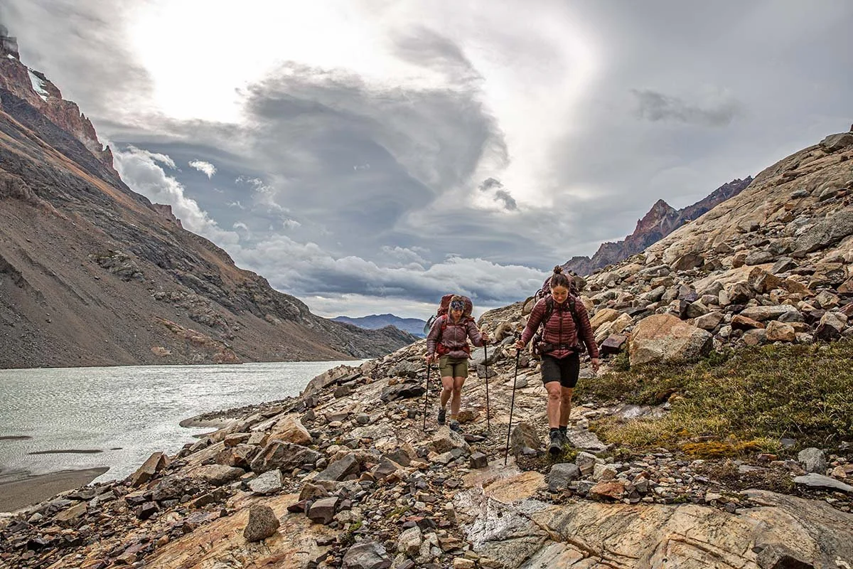 Two hikers with backpacks and trekking poles walking along a rocky trail beside a lake, surrounded by mountains and overcast sky.