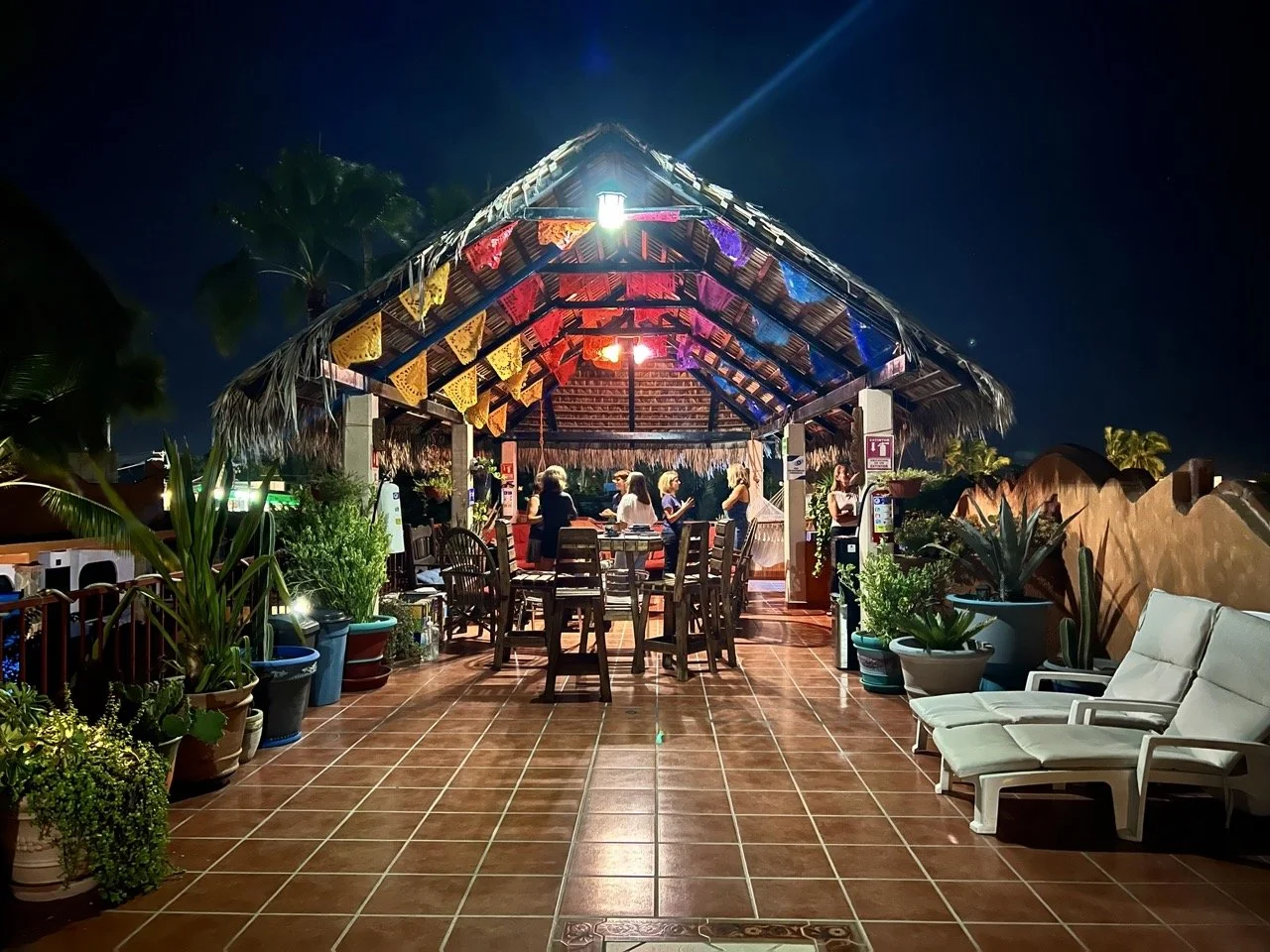 Night scene at an outdoor restaurant or bar with string lights, tropical plants, and a thatched roof. People are standing and sitting around tables, with chairs and lounge chairs nearby. The atmosphere is lively and relaxed.