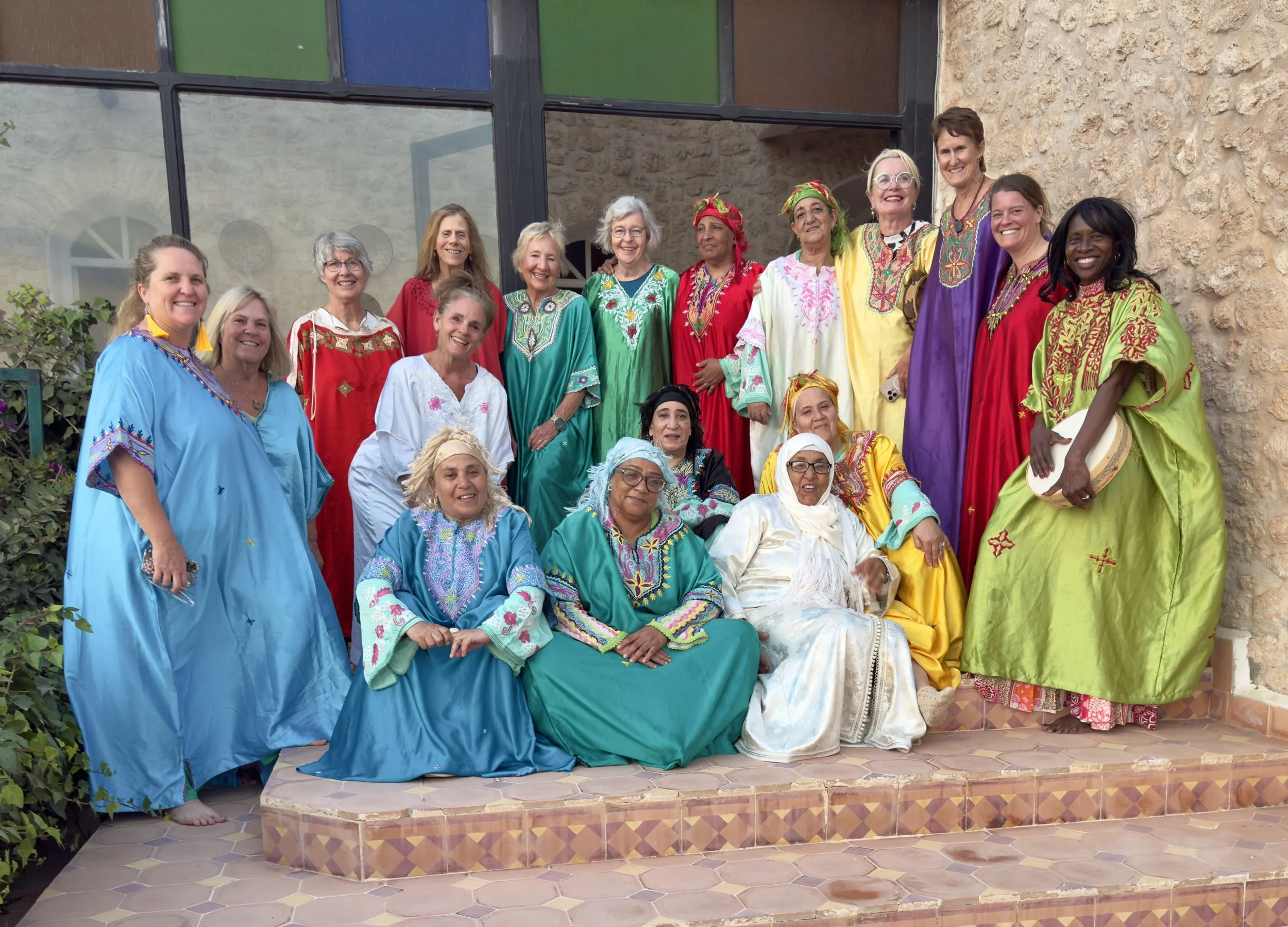 Group of women wearing colorful traditional dresses and costumes standing and sitting on steps outside a building with glass windows and stone wall.
