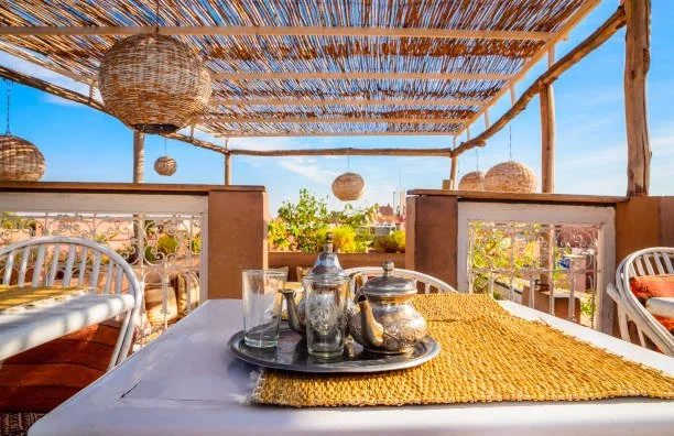 Outdoor patio with a white dining table, silver teapot and cups, yellow table runner, wicker hanging lights, and a bamboo roof, overlooking a cityscape under a blue sky.