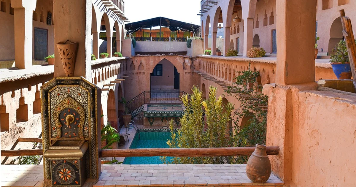 A traditional Moroccan courtyard with a small swimming pool, colorful ceramic tiles, potted plants, and arched openings in pinkish walls, featuring Moroccan decorative elements.