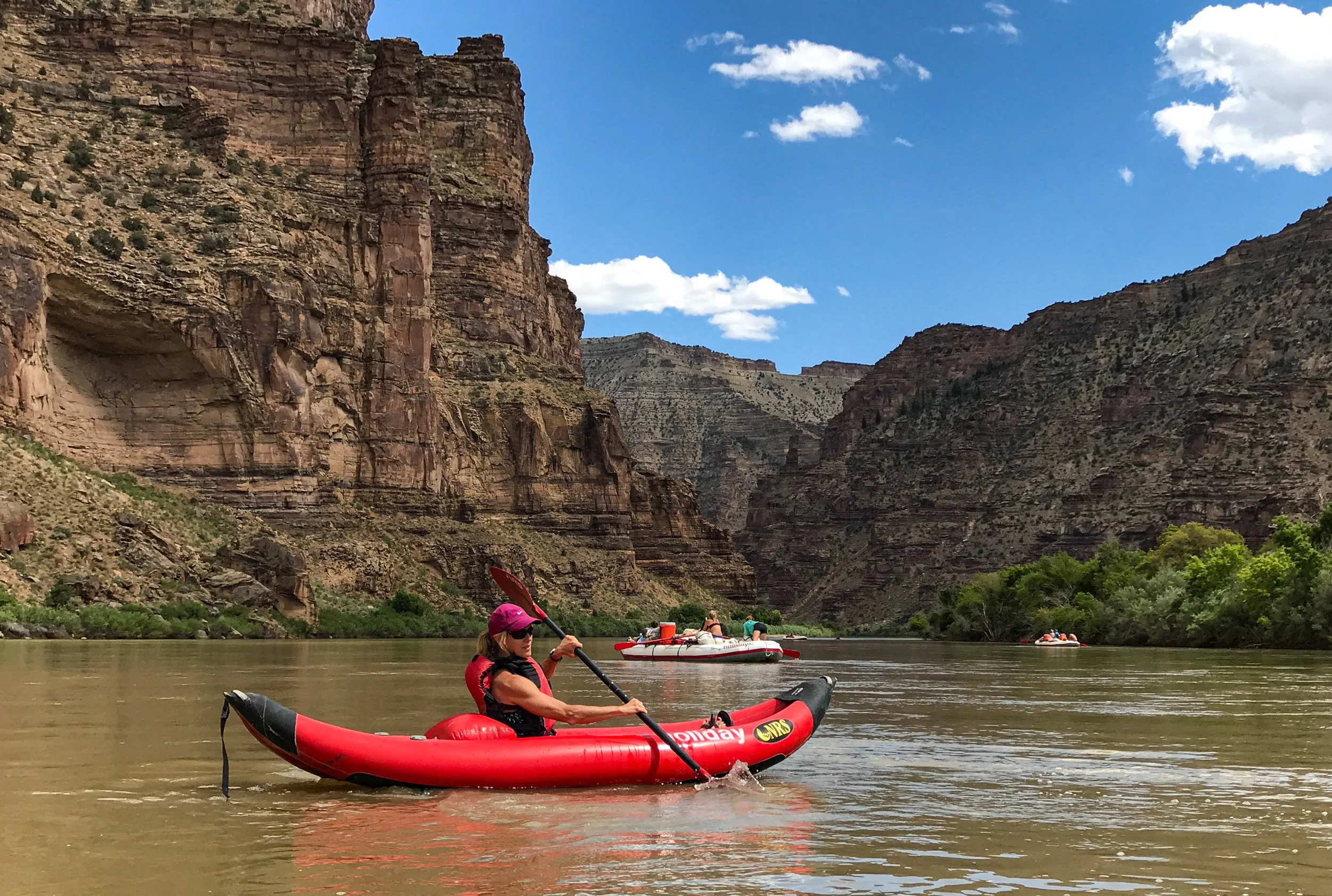 Woman kayaking on a river surrounded by tall canyon cliffs, with other boats in the background and a partly cloudy sky.