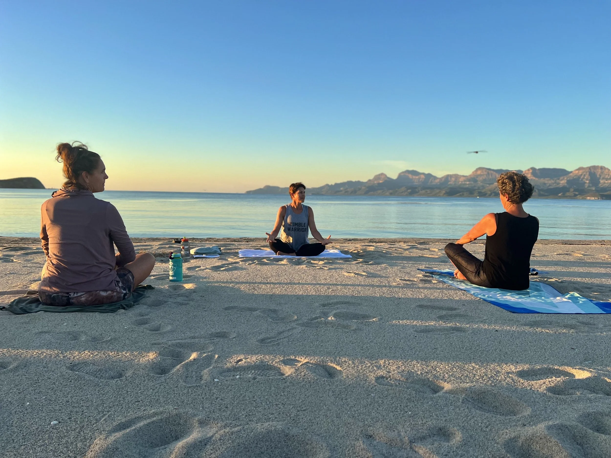 Three people practicing yoga on a sandy beach during sunrise or sunset, with mountains across calm water in the background.
