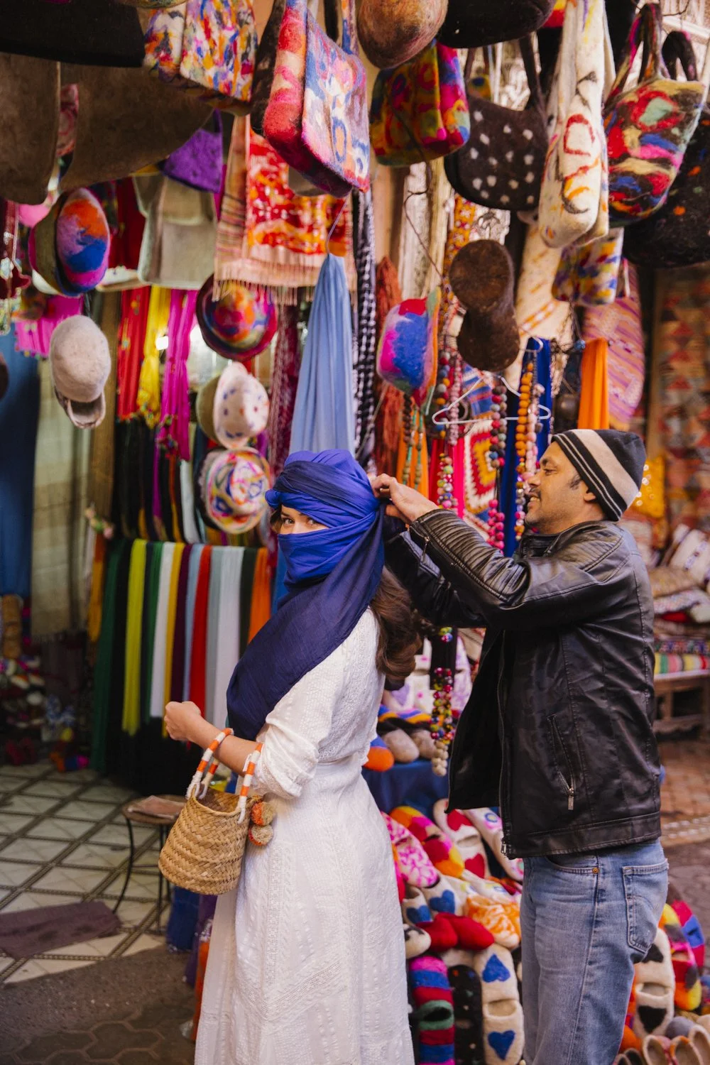 A woman in a white dress and blue headscarf with a basket is shopping at a colorful market stall with hats, scarves, and accessories. A man helps adjust her headscarf, both smiling amid vibrant textiles and merchandise.