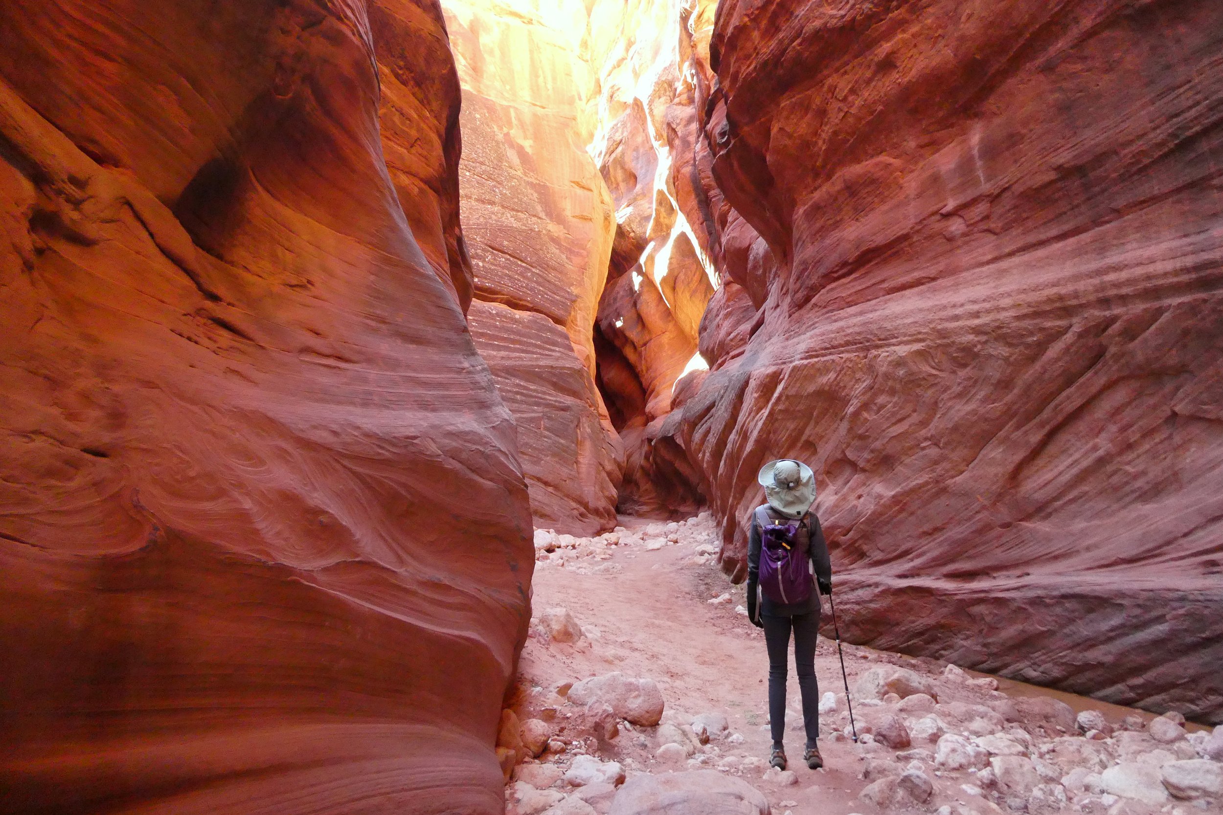 Hiker with a purple backpack and walking stick exploring a narrow, winding slot canyon with reddish-orange layered rock walls.