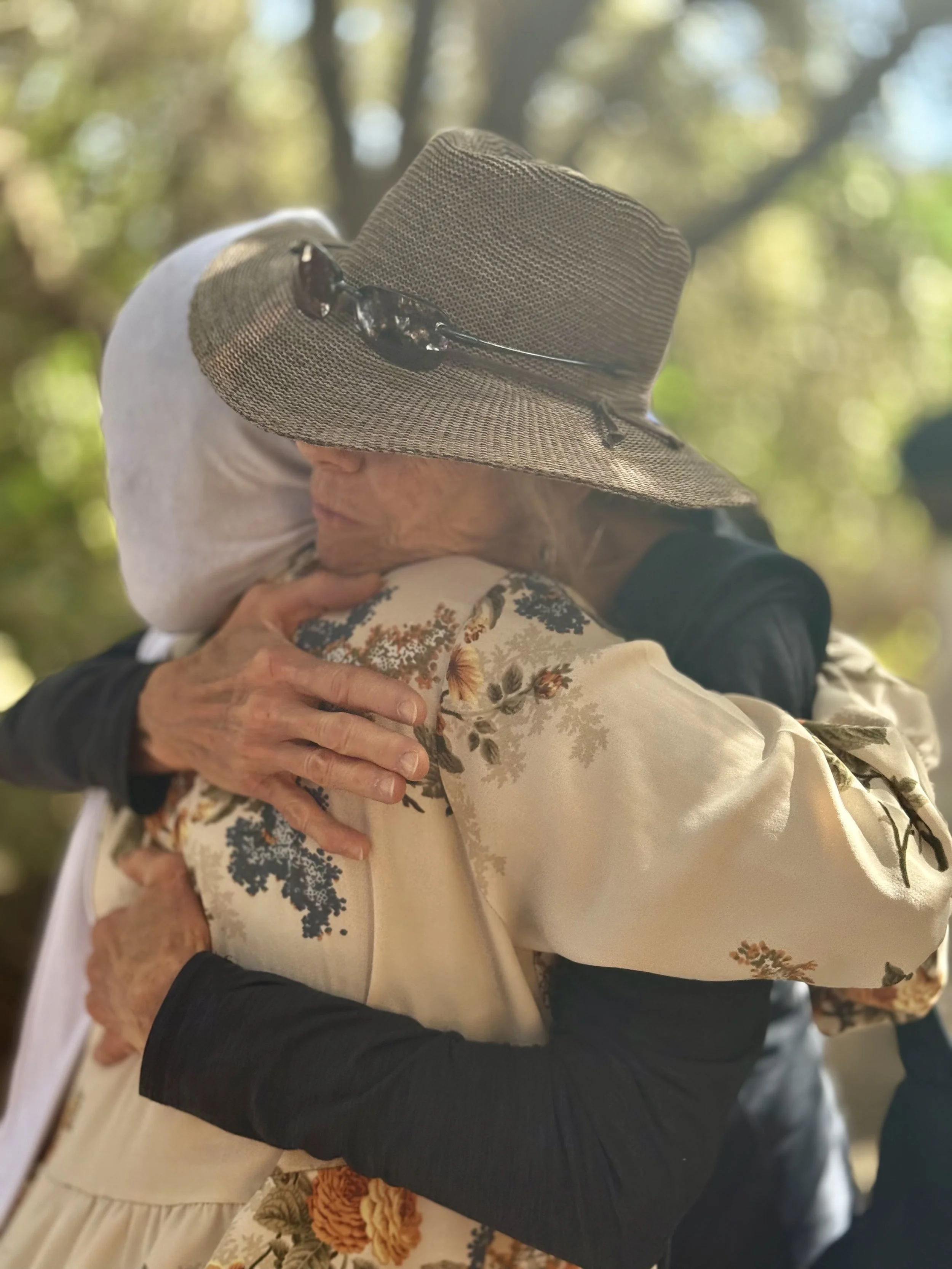 Two elderly women hugging outdoors beneath a large sun hat worn by one of them, with a blurred background of green trees.