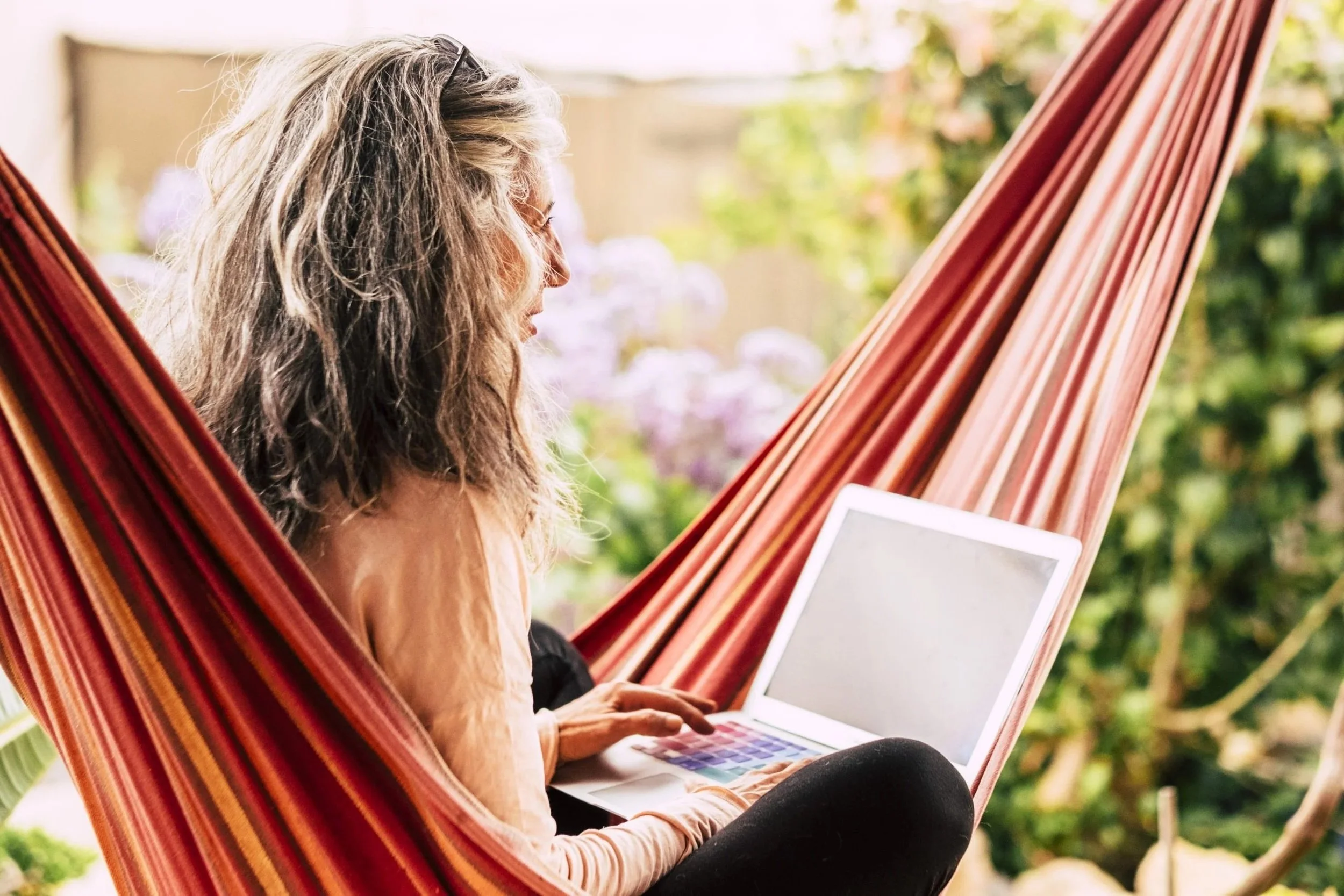 A woman with long, wavy gray hair sitting in a hammock outdoors, using a laptop.