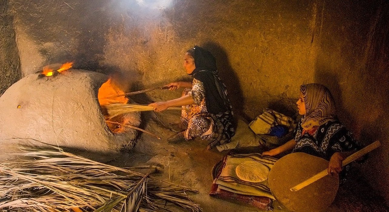Two women wearing patterned headscarves and traditional clothing in a cave or underground setting. One woman is tending a fire inside a rounded clay oven, while the other is sitting nearby holding a wooden paddle or stick. The scene appears to be traditional or artisanal.